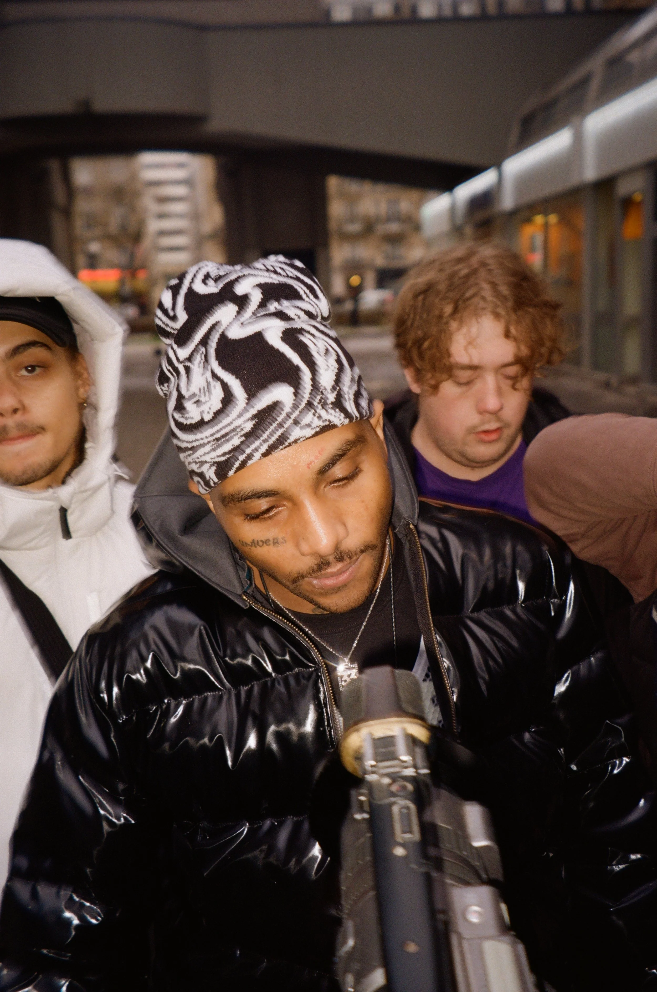 Group of young men standing close together under an overpass at night, with one holding a gun aimed at the camera, in an urban setting.