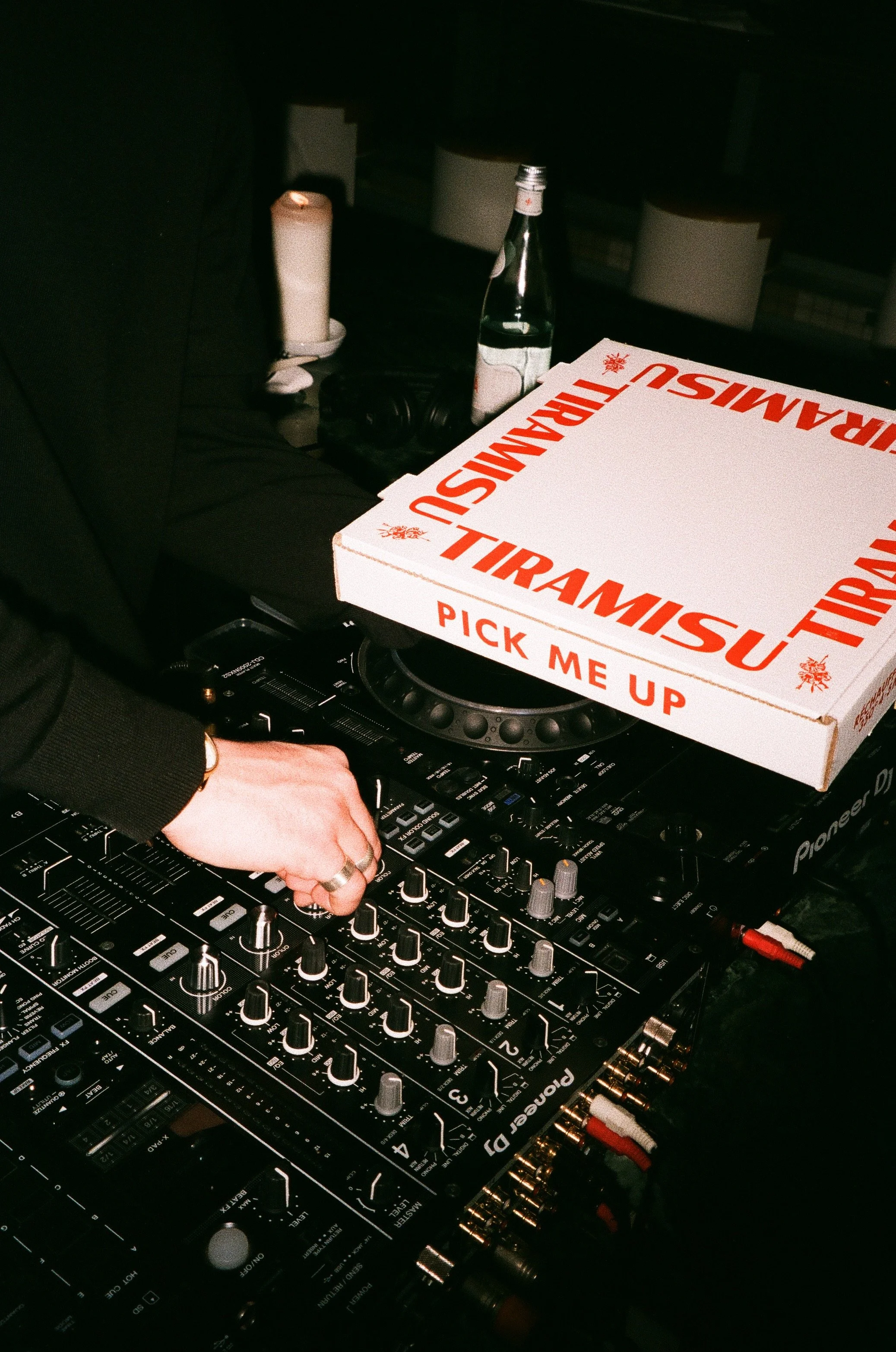 A DJ's hand adjusting controls on a Pioneer DJ mixer, with a box of Tiramisu pizza, a green glass bottle, a candle, and headphones on the table.