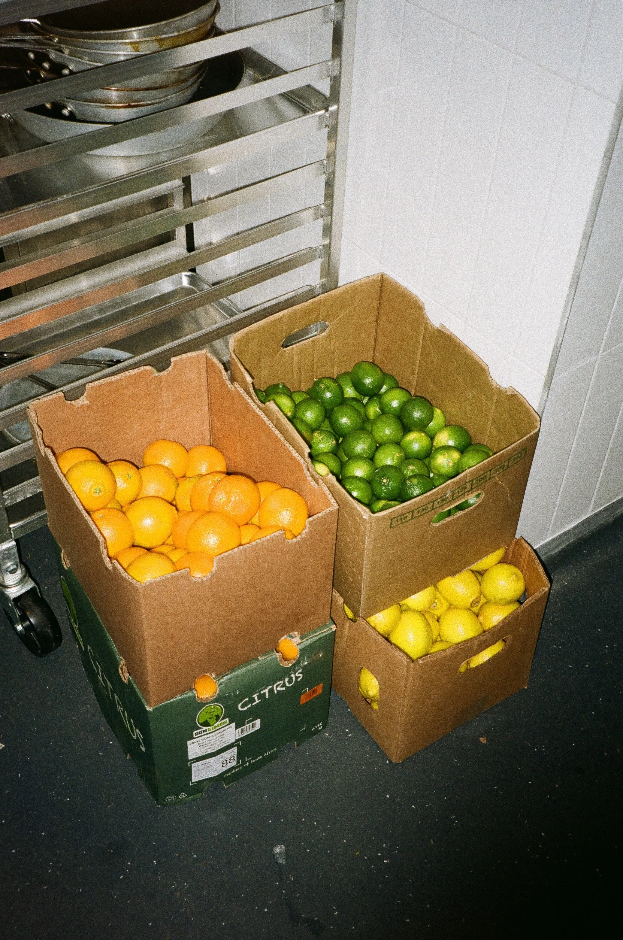 Three cardboard boxes containing oranges and limes near a metal shelf in a kitchen.