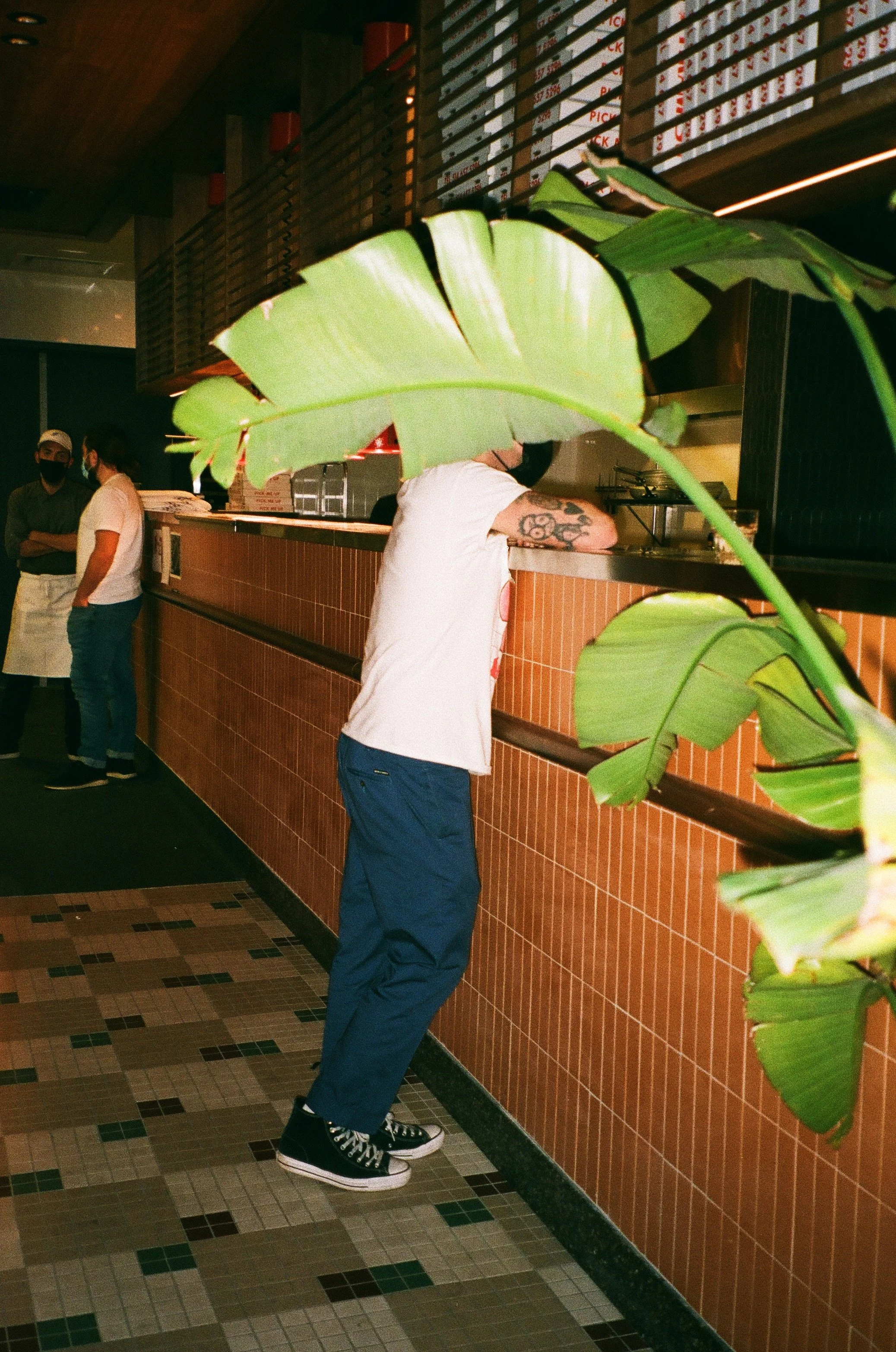 Person with arm tattoos leaning on a counter inside a restaurant, partially obscured by large green leaves in the foreground, with two other people standing in the background wearing masks and casual outfits.