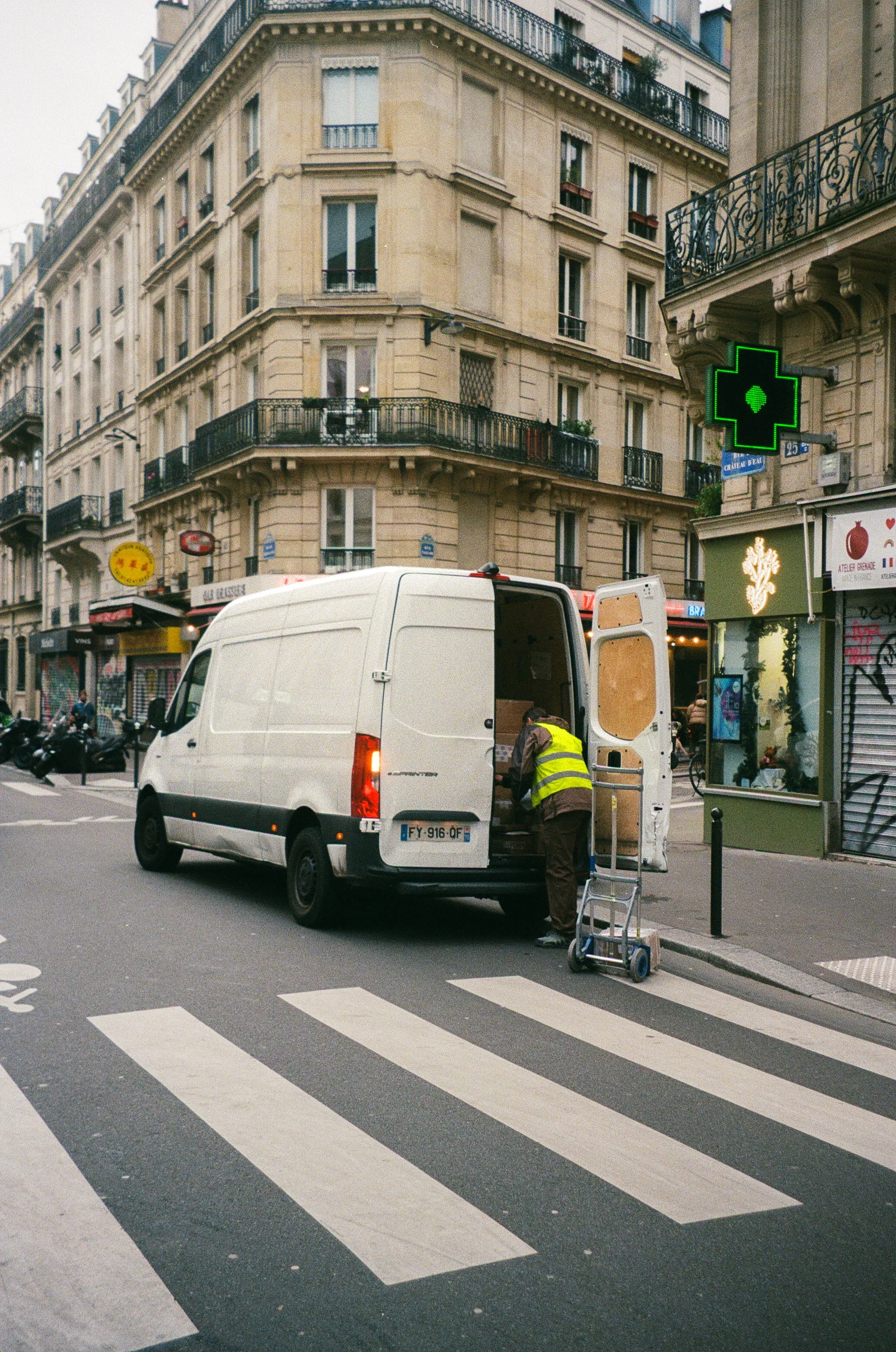 A worker in a yellow safety vest loading goods into a white delivery van parked on a city street with crosswalk.