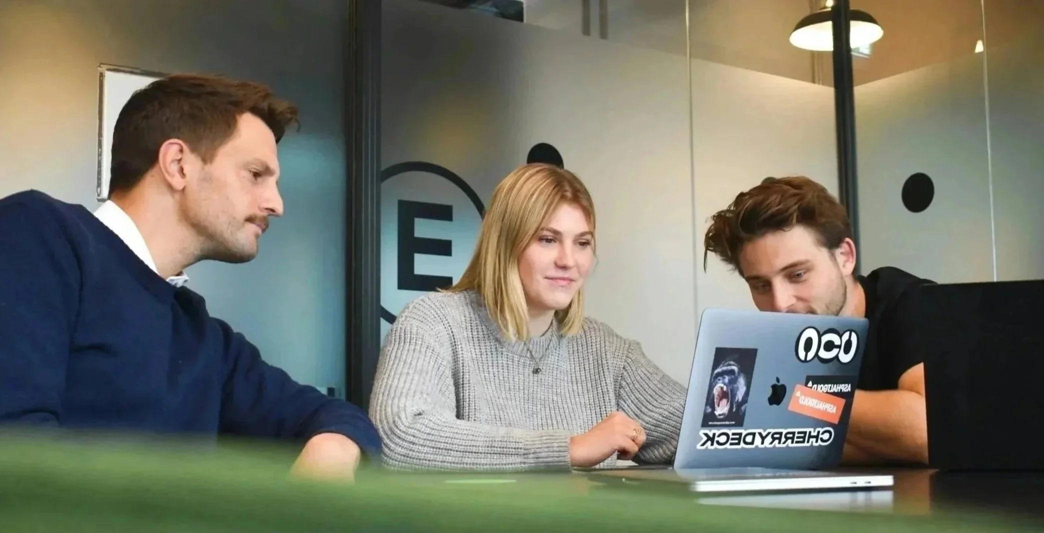 Three people sitting at a table looking at a laptop in an office.