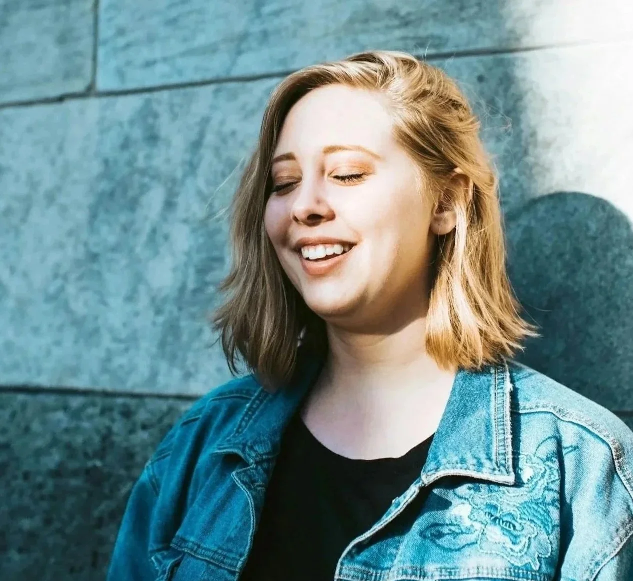 A young woman with shoulder-length blond hair smiling with her eyes closed, leaning against a gray stone wall partly illuminated by sunlight.