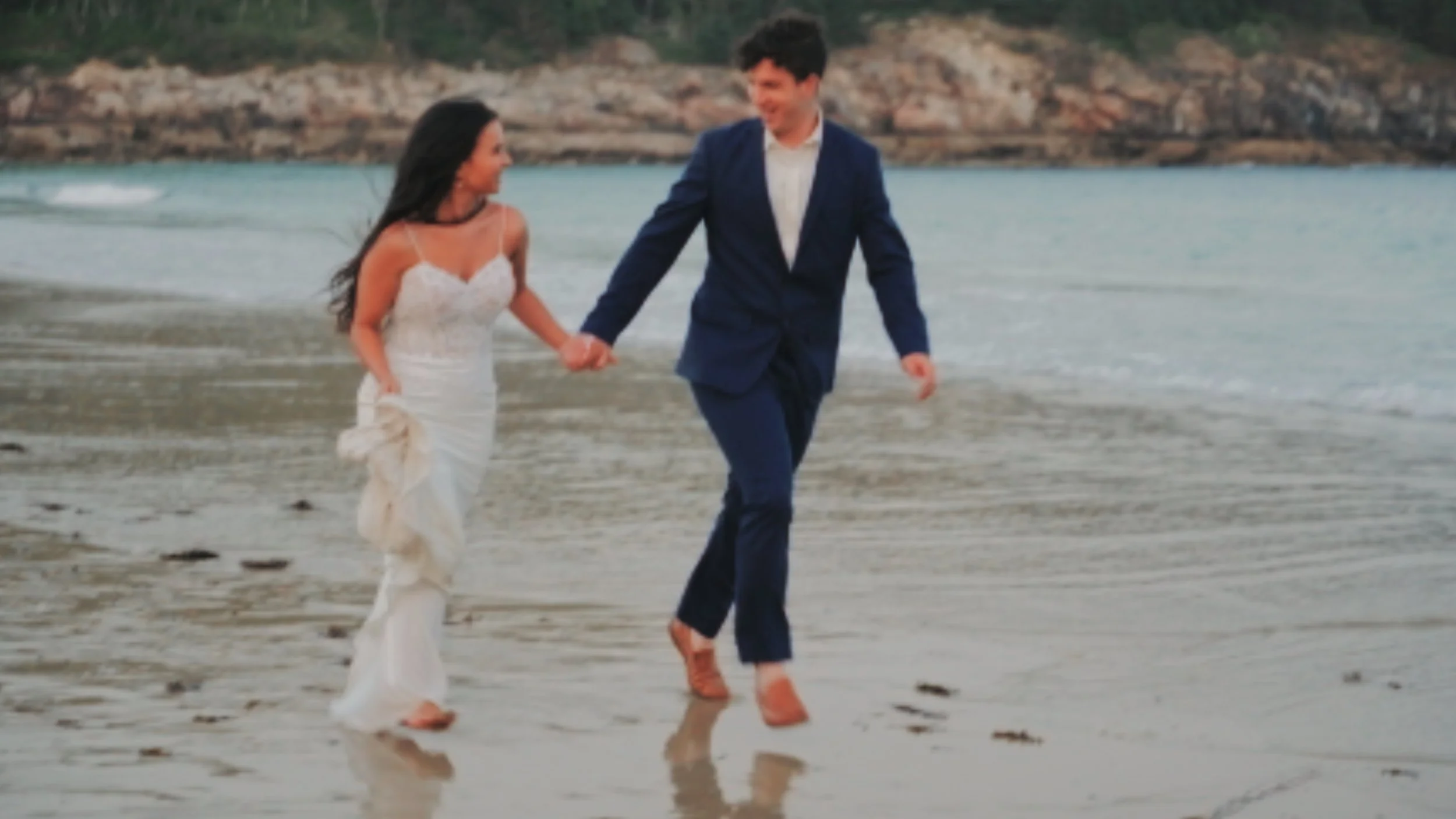 A couple walking hand-in-hand on the beach, with the woman in a white dress and the man in a blue suit, holding hands and smiling, with water and rocky cliffs in the background.