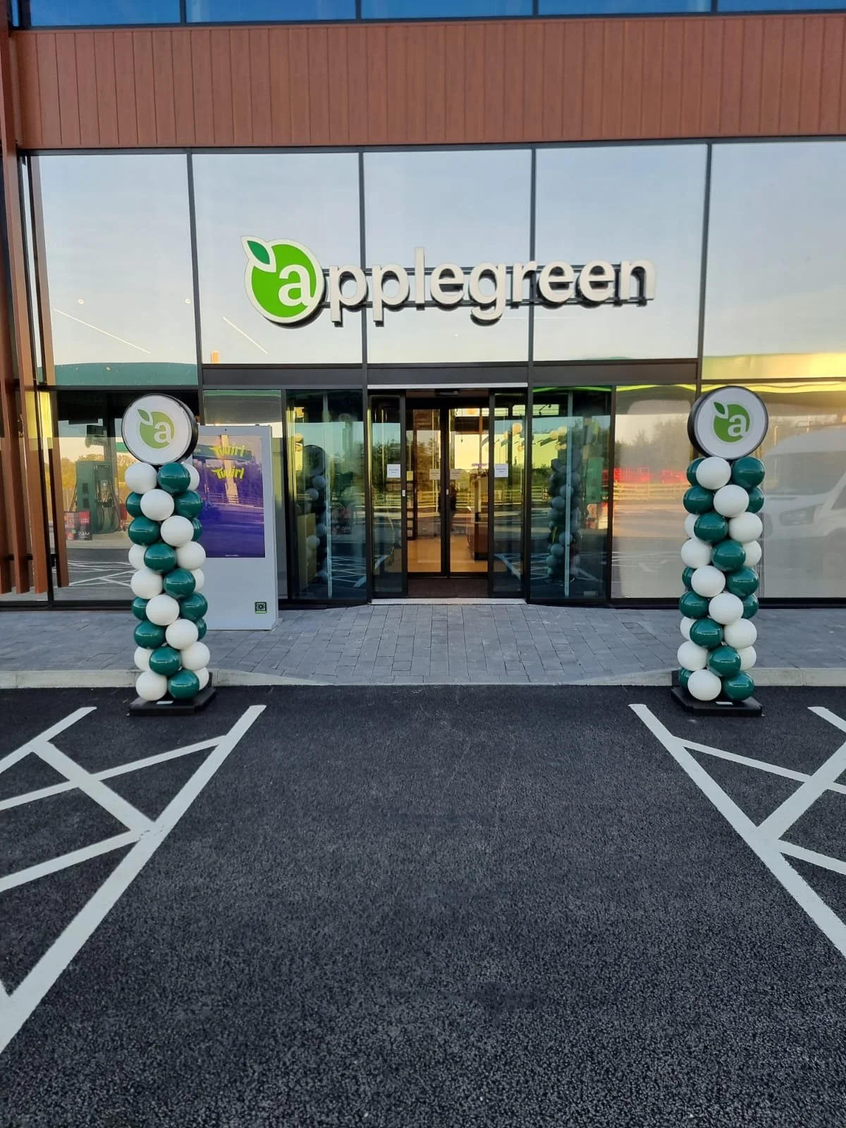 Exterior view of a building with a glass façade and the sign 'aplegreen' on top. There are two balloon columns decorated with green and white balloons on either side of the entrance. The entrance has glass doors, and a parking lot with marked parking spaces is in front.