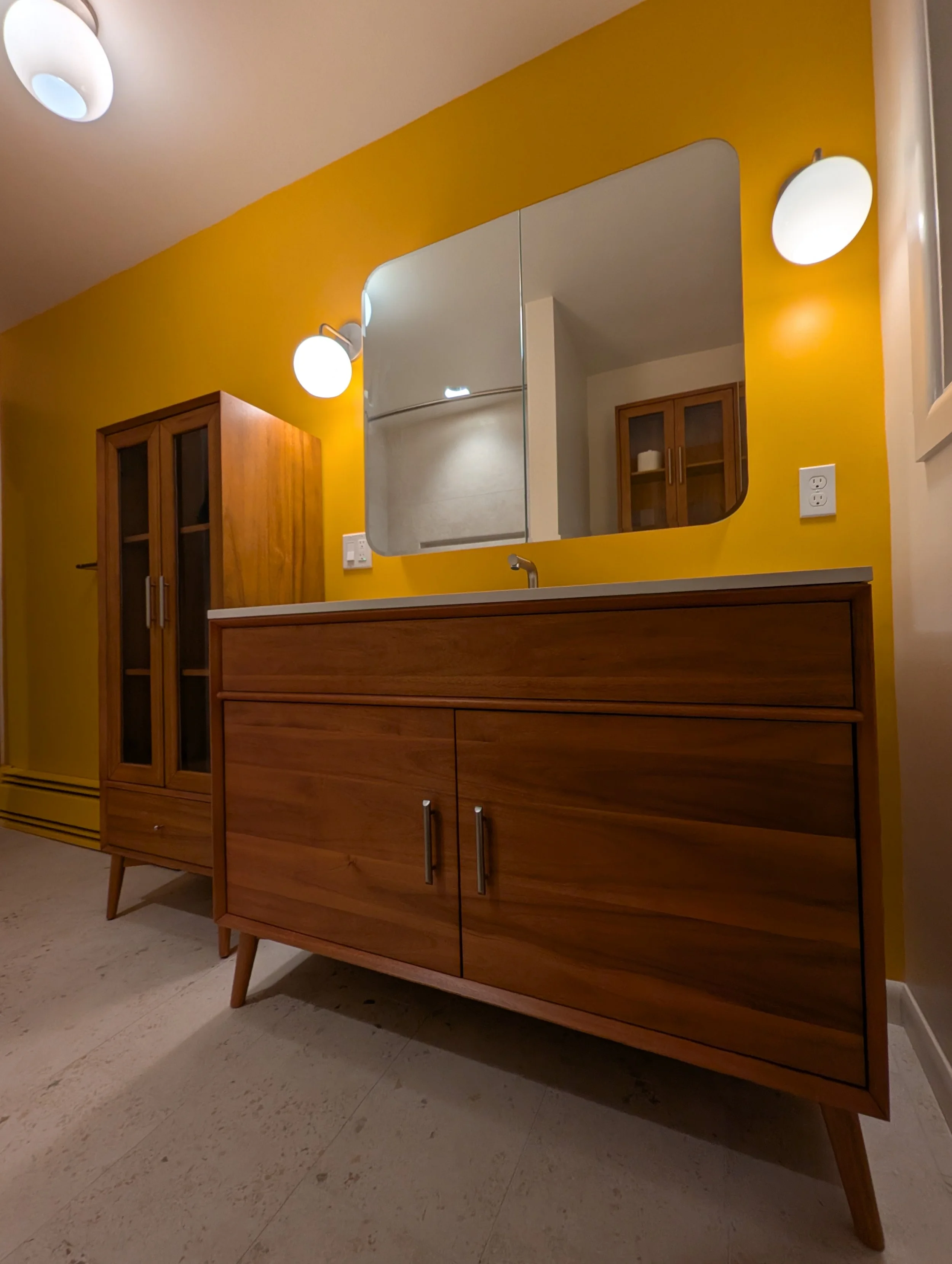 A remodeled wooden cabinet with two doors and metal handles, placed on four angled legs, against a yellow wall with a large rectangular mirror and two white wall sconces.