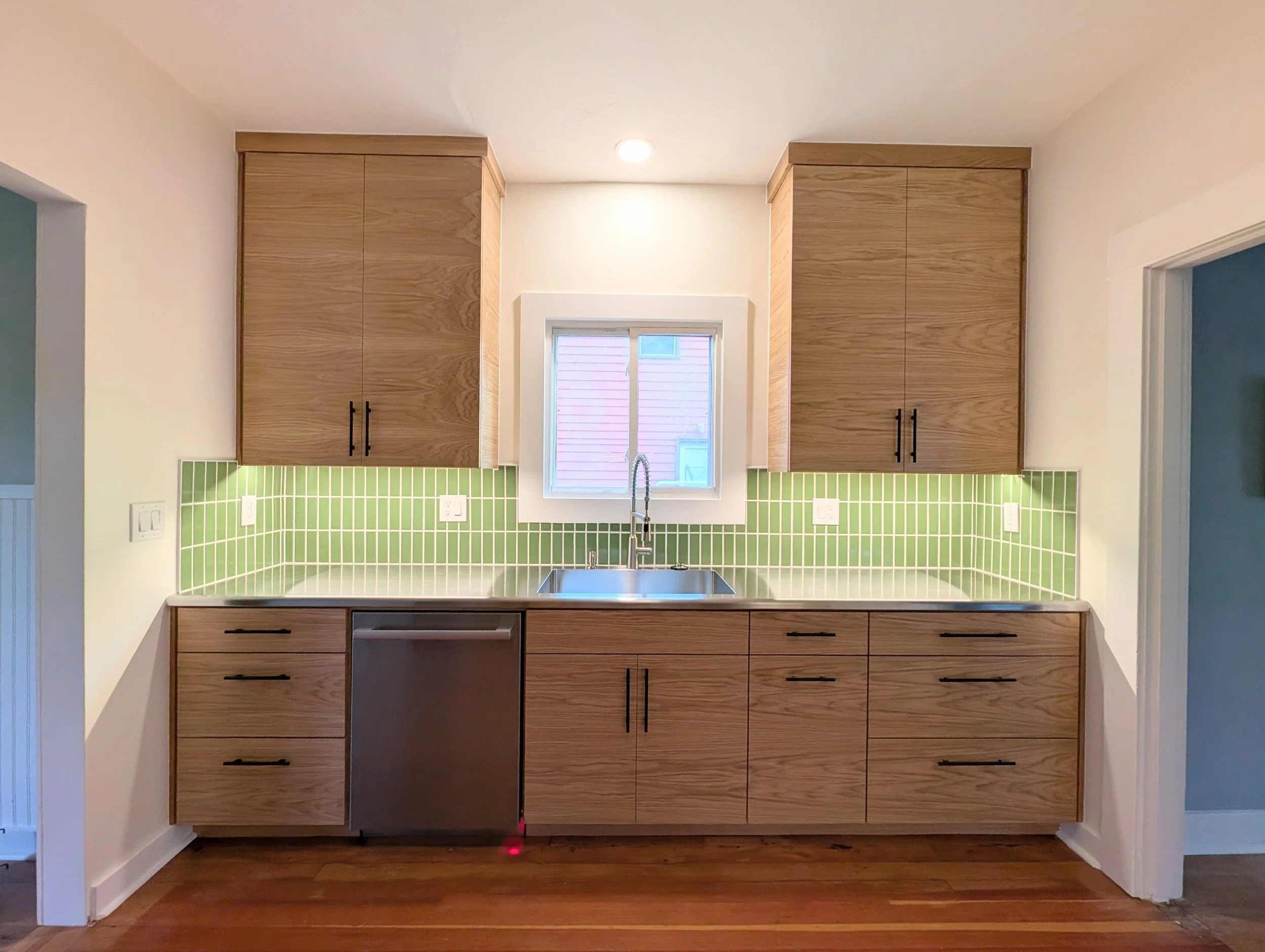 A remodeled kitchen with wooden cabinets, a green tiled backsplash, and a window above the sink.