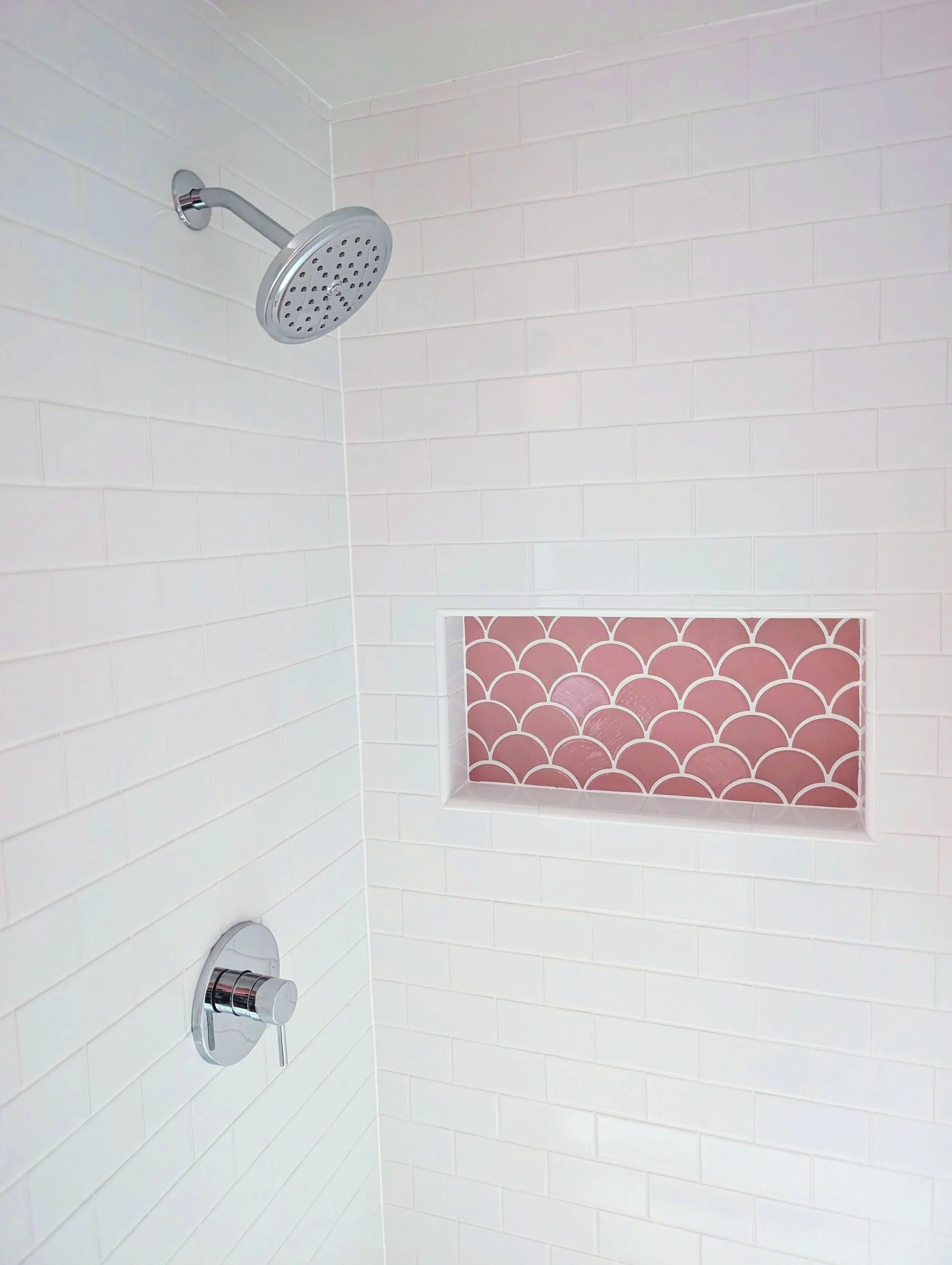 Shower with white tiled walls, a round showerhead, a control handle, and a pink decorative niche with a pattern.