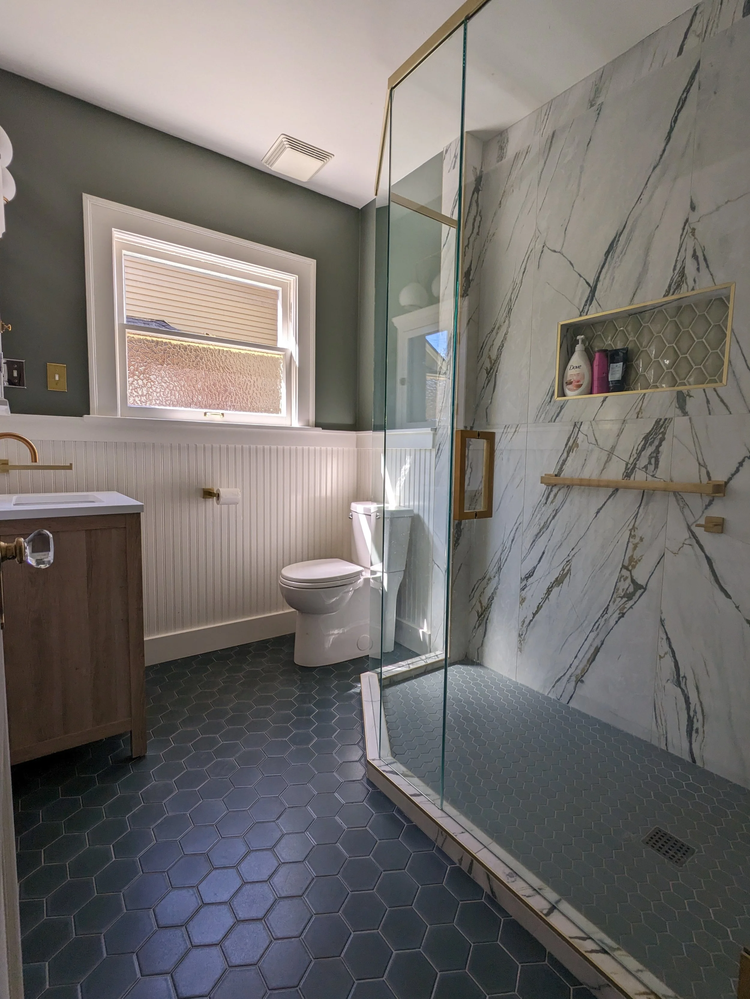 A luxury bathroom remodel with a gray and white color scheme, featuring a toilet, a vanity with a sink, a window with blinds, and a walk-in shower with marble walls and gray hexagonal tiles.
