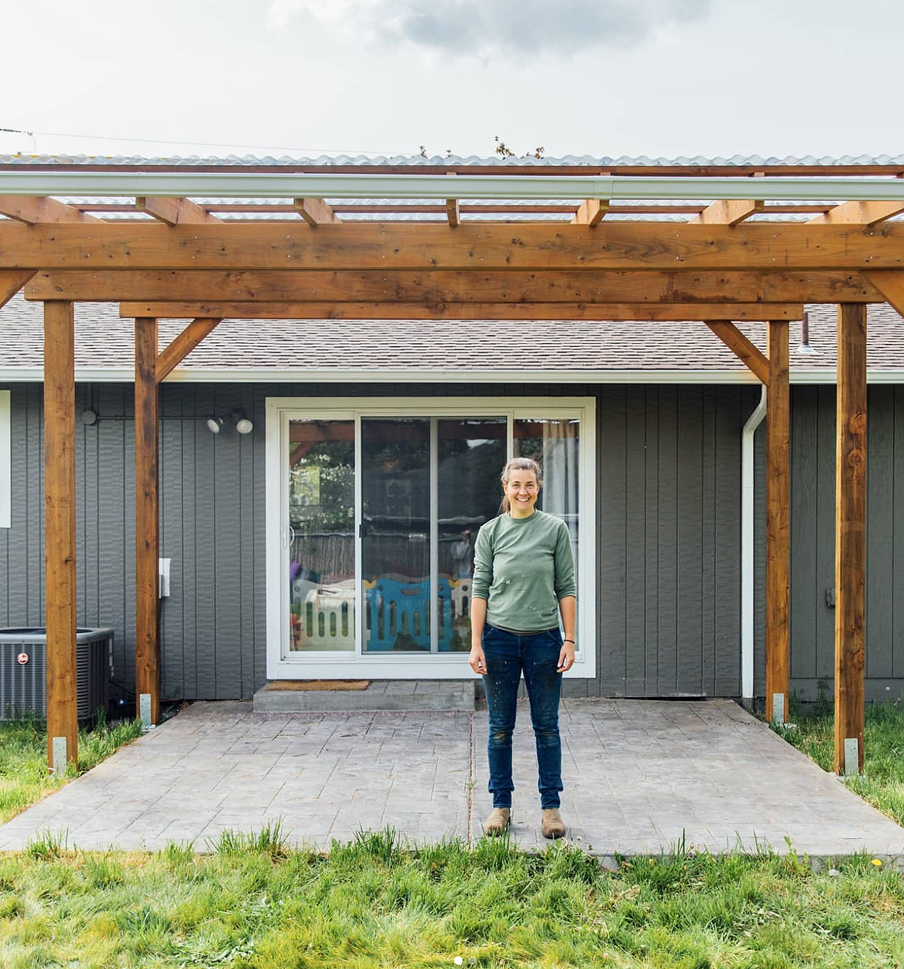 Ann Lawson standing on a paved patio in front of a sliding glass door. The patio is covered by a wooden pergola under construction, with beams and support posts visible. The house has dark gray siding and a shingled roof.