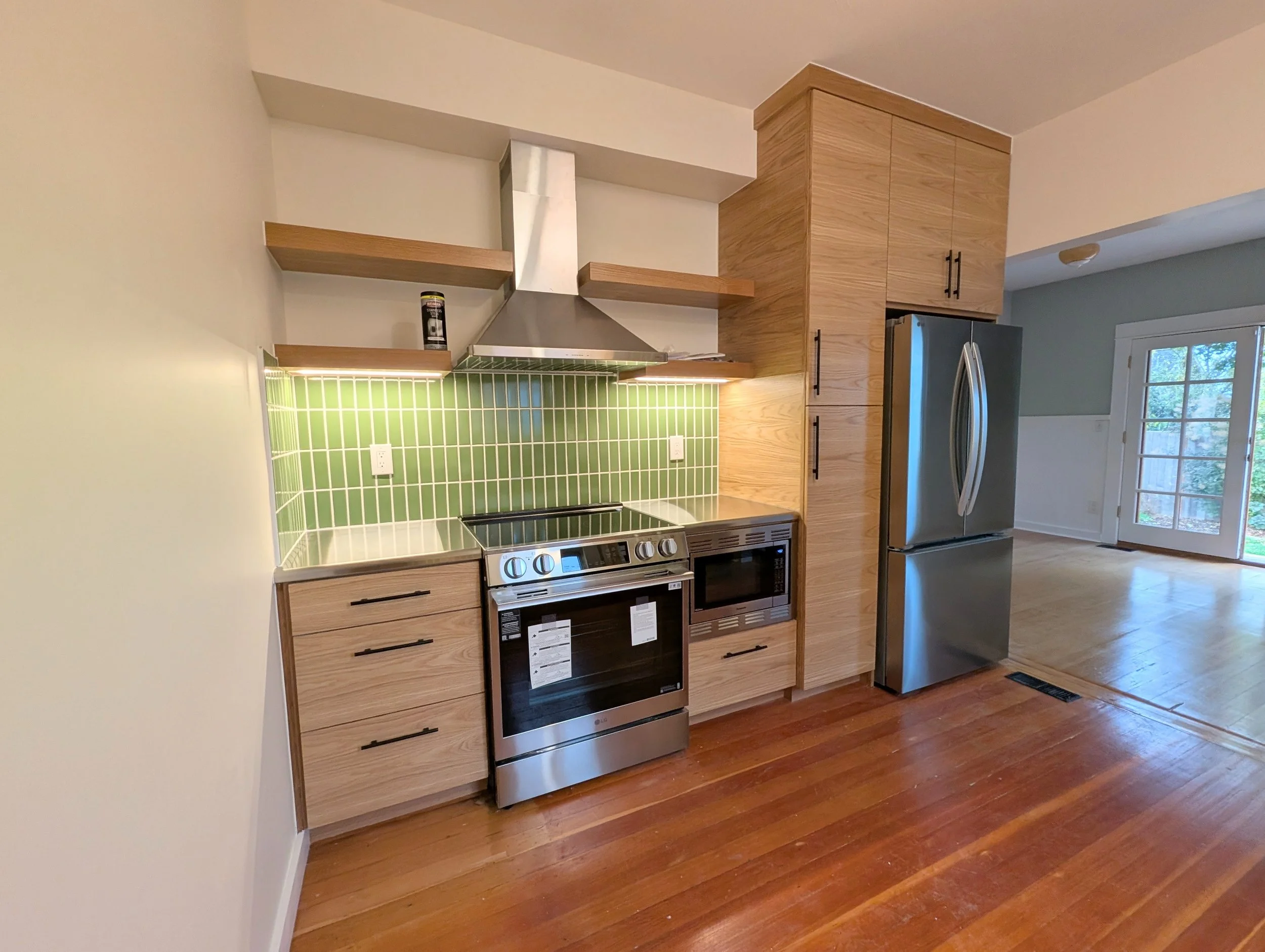 Modern remodeled kitchen with wooden cabinets, stainless steel appliances, green tile backsplash, and open shelving, with hardwood flooring and a view of the living area with a sliding glass door.