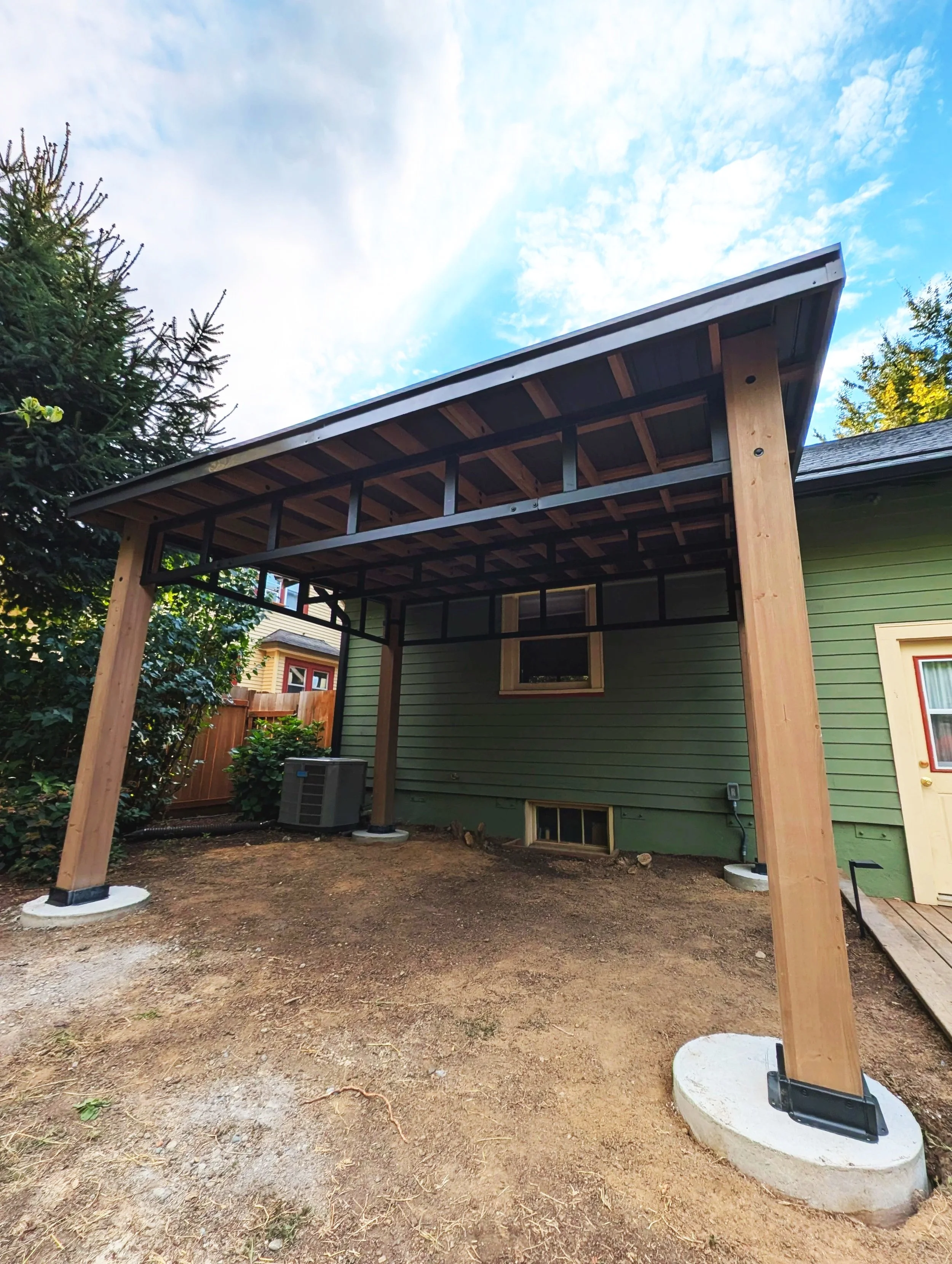 Backyard with new wooden porch structure under construction, green house in background, dirt ground, blue sky with some clouds.