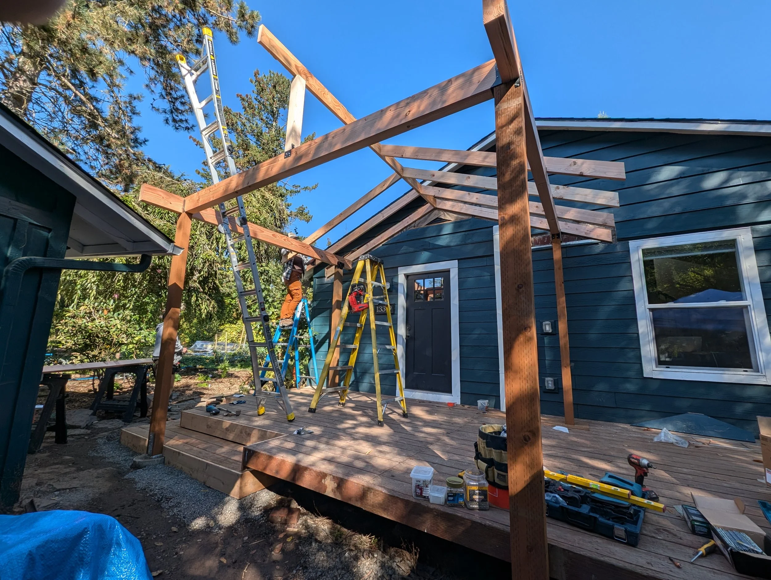 Construction of a wooden deck attached to blue house, with workers on ladders installing roof beams under clear blue sky.