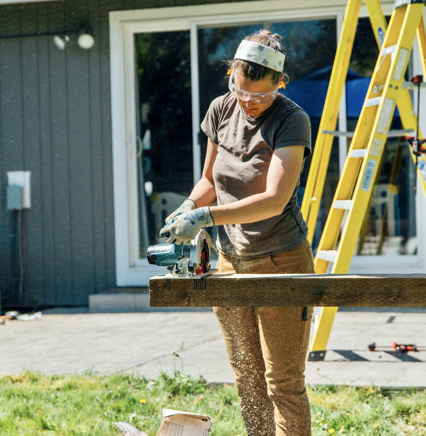 Ann Lawson wearing safety glasses, gloves, and a headband operating a handheld circular saw on a piece of wood outdoors near a gray house with a sliding glass door, a yellow ladder, and a blue canopy in the background.