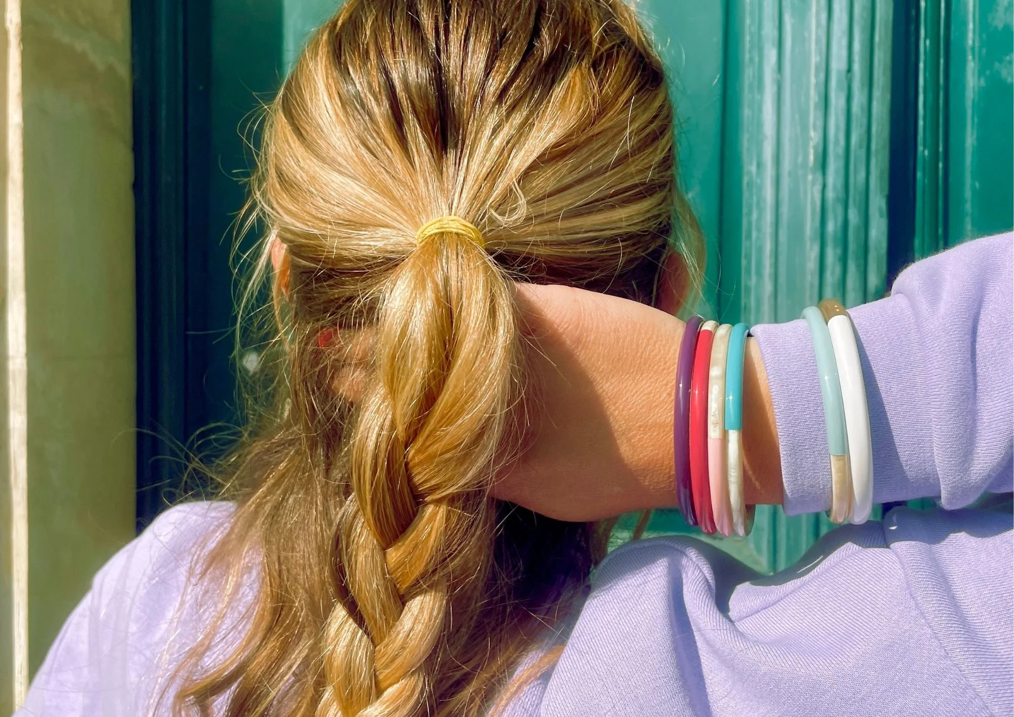 Femme aux cheveux blonds attachés en queue de cheval, portant plusieurs bracelets colorés, assise devant une porte en bois vert. vue de dos.