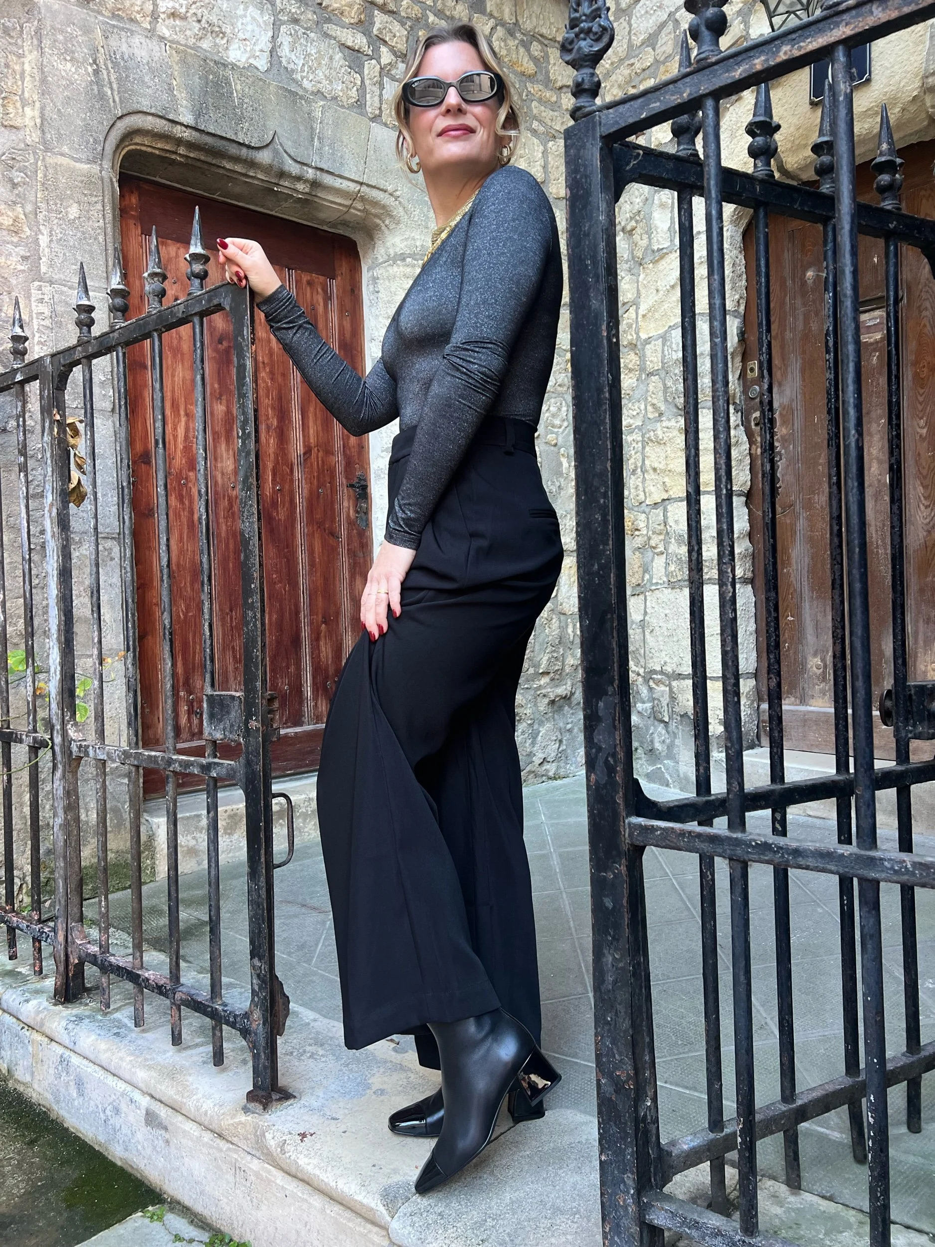 Femme élégante portant des vêtements sombres, lunettes de soleil, chaussures à talons et haut moulant, posant sur un passage en pierre devant une porte en bois entourée d'un mur en pierre et une clôture en fer forgé.