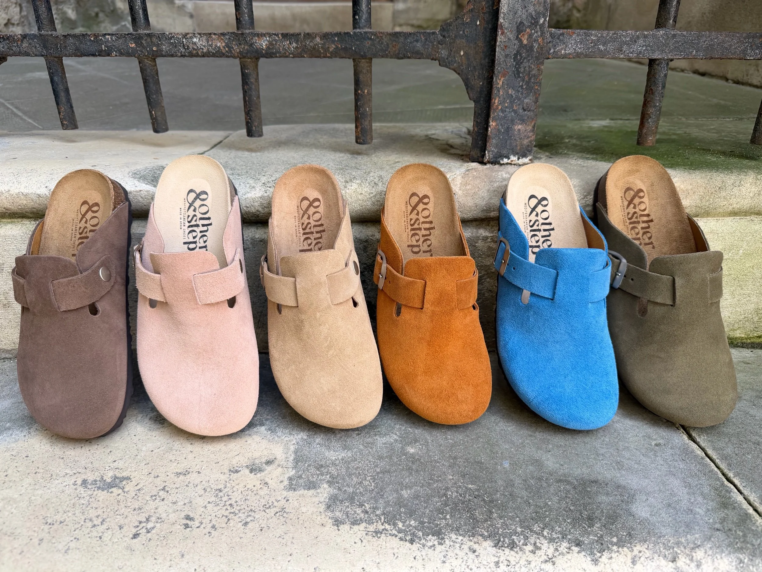 Une rangée de cinq paires de sabots en cuir ou en suède de différentes couleurs, disposés sur un trottoir en pierre sous une barrière en fer forgé.