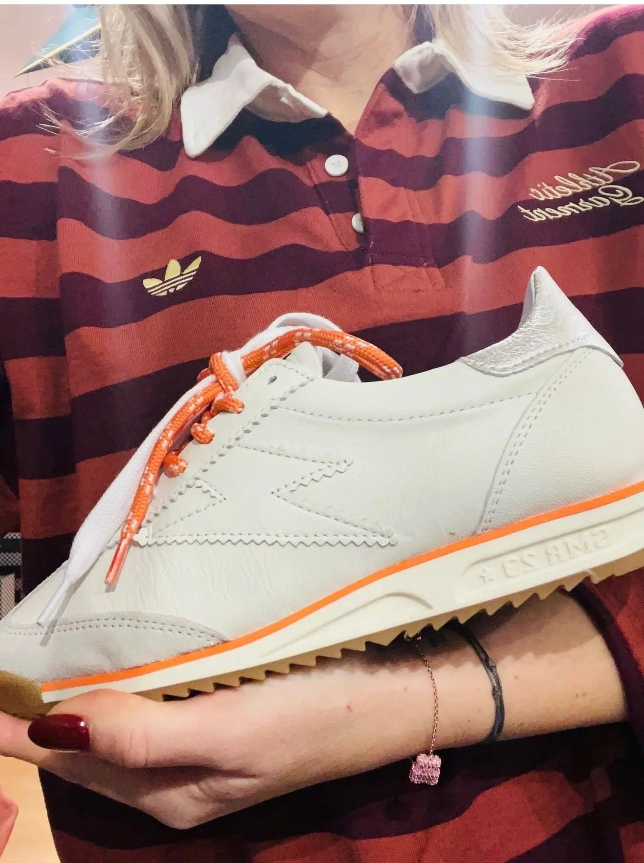 Une personne portant une chemise à rayures rouges et noires avec un col blanc, tenant une chaussure blanche avec des lacets orange. La chaussure est en gros plan.