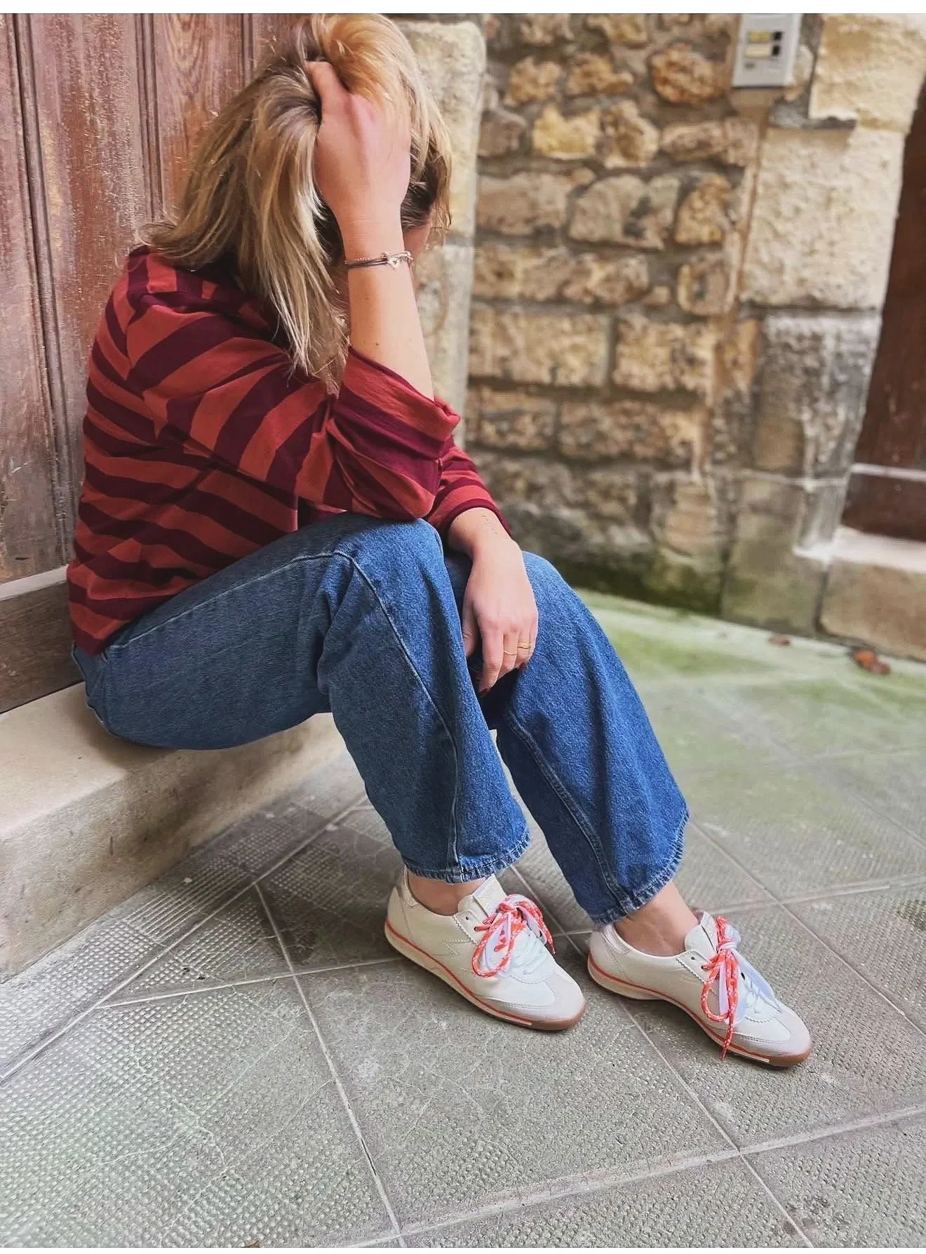 Une femme assise par terre contre un mur en pierre, avec sa main sur la tête, portant un sweat à rayures rouges et marron, un jean ample et des baskets blanches avec des lacets rouges.
