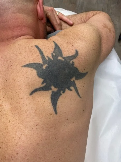 Close-up of a man's shoulder with a large black tribal-style tattoo, resting on a white pillow or cloth.