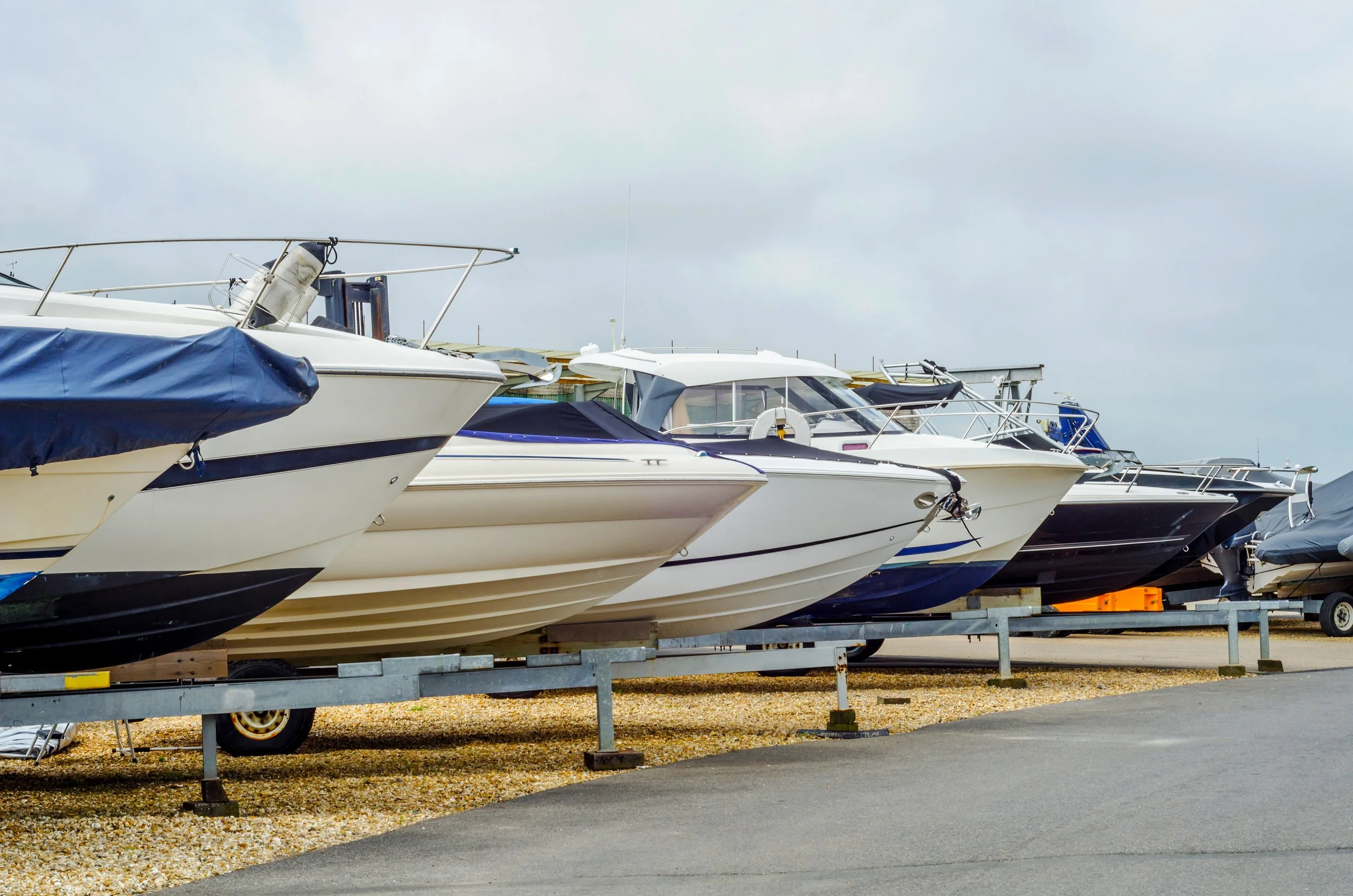 Row of boats on trailers in a marina, parked on gravel next to a paved area under an overcast sky.