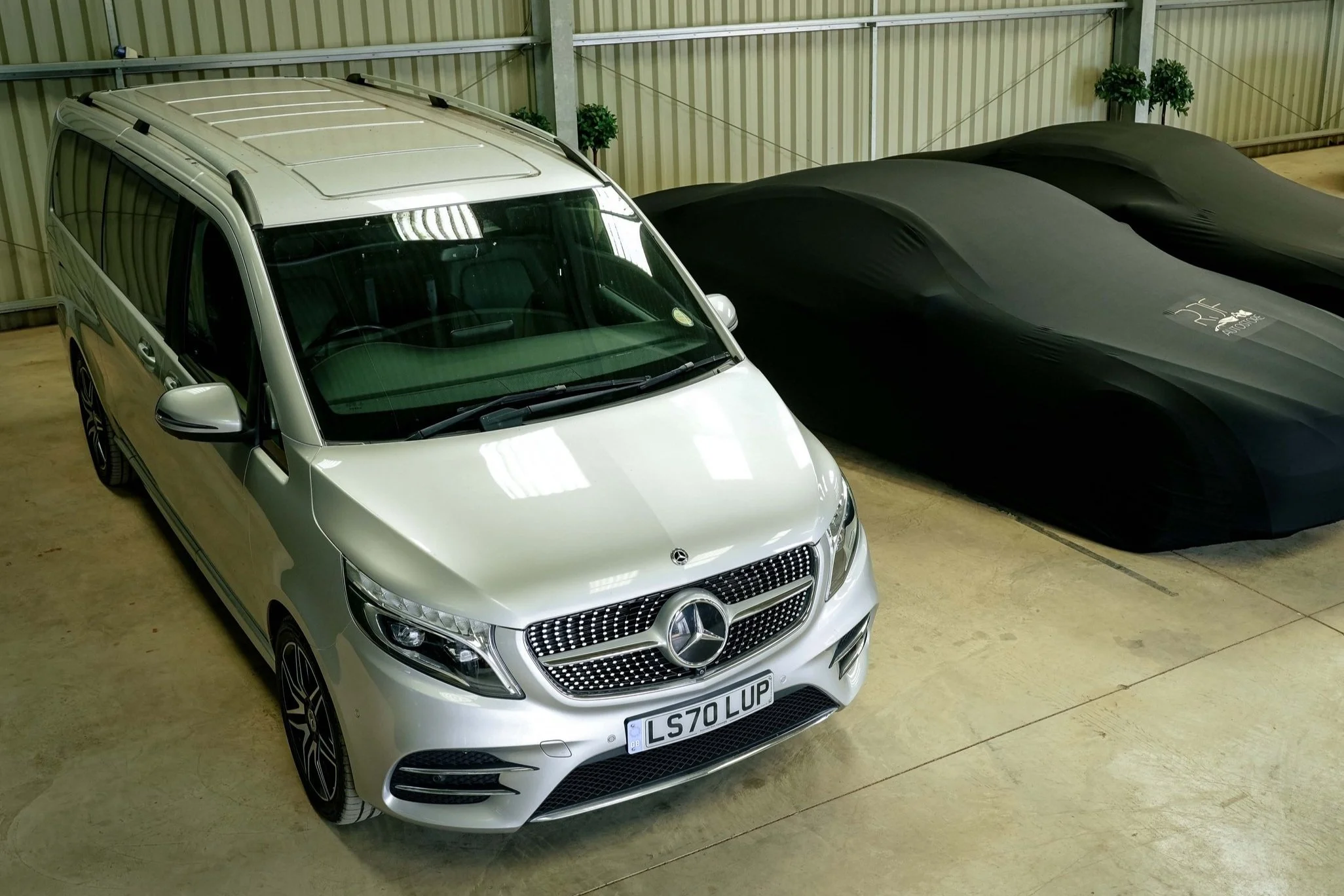 Silver Mercedes-Benz van parked in a garage next to two covered vehicles.