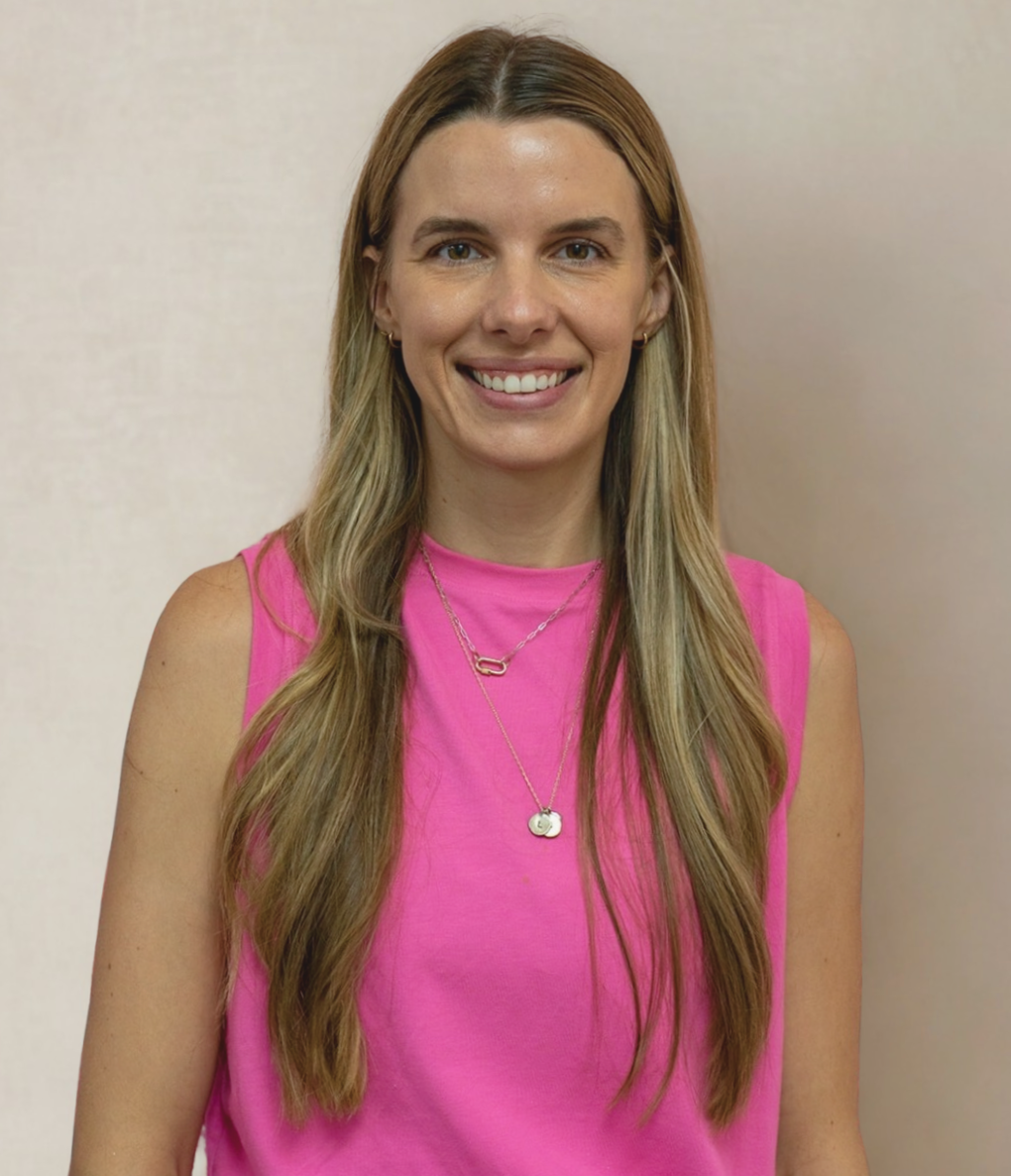 A woman with long blonde hair wearing a pink sleeveless top and layered necklaces, smiling at the camera against a plain beige wall.