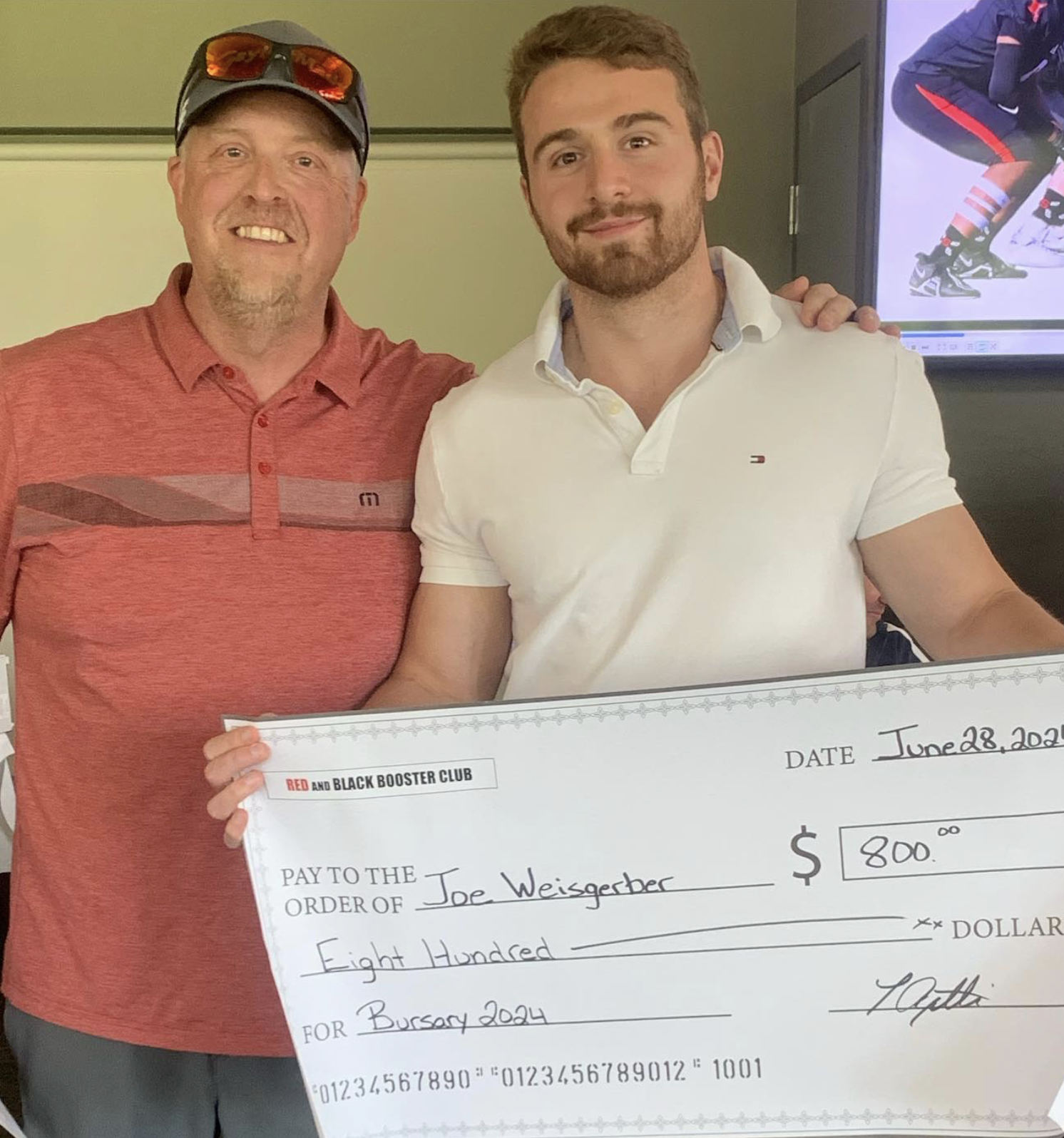 Two men smiling and holding an oversized check made out to Joe Weiserber for $800, dated June 28, 2024, with a background showing a television displaying a hockey player. One man wears a red shirt and baseball cap, and the other wears a white shirt.