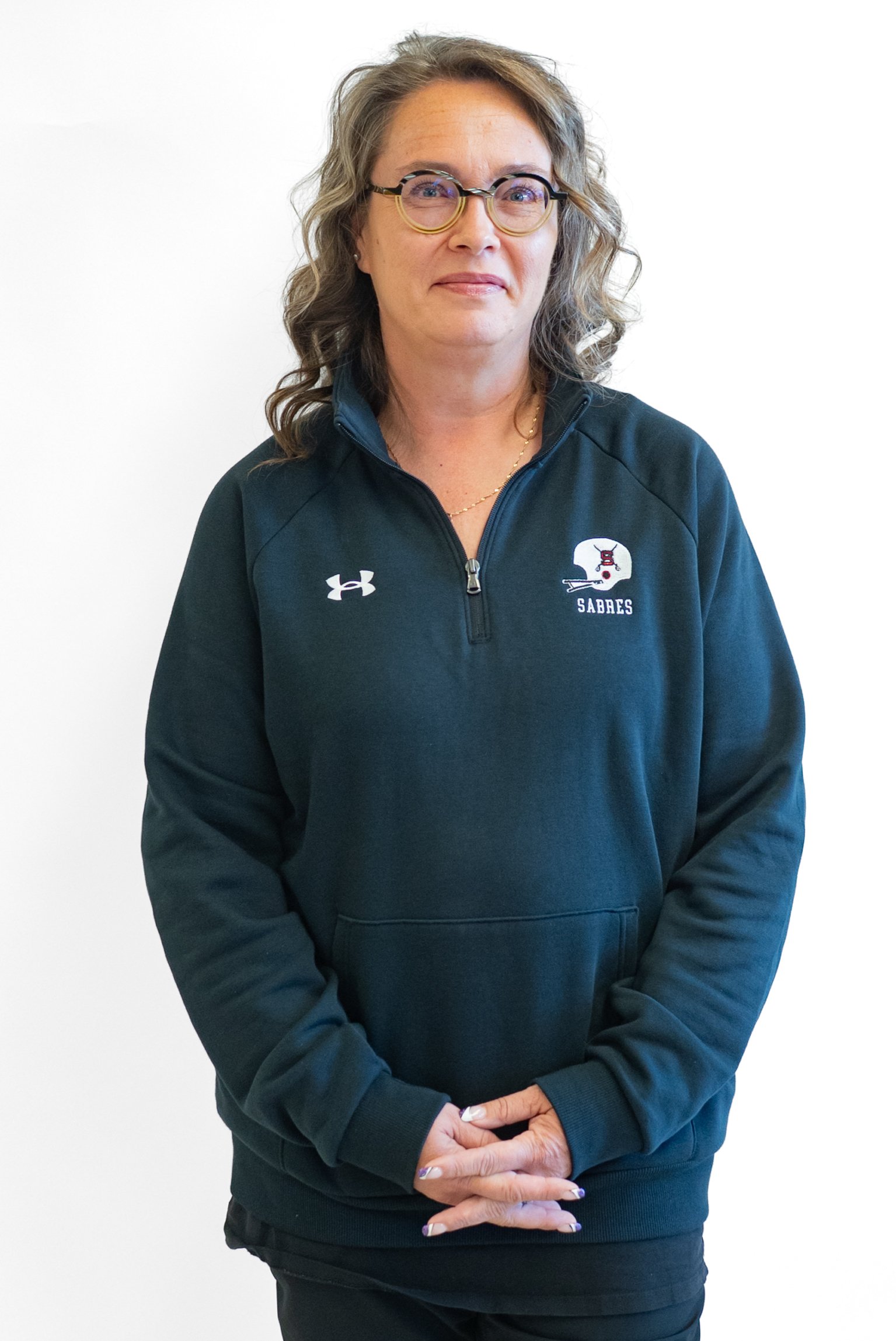A woman with glasses and hoop earrings smiling, wearing a black jacket with 'SAL FOOTBALL' on it, standing against a brick wall.