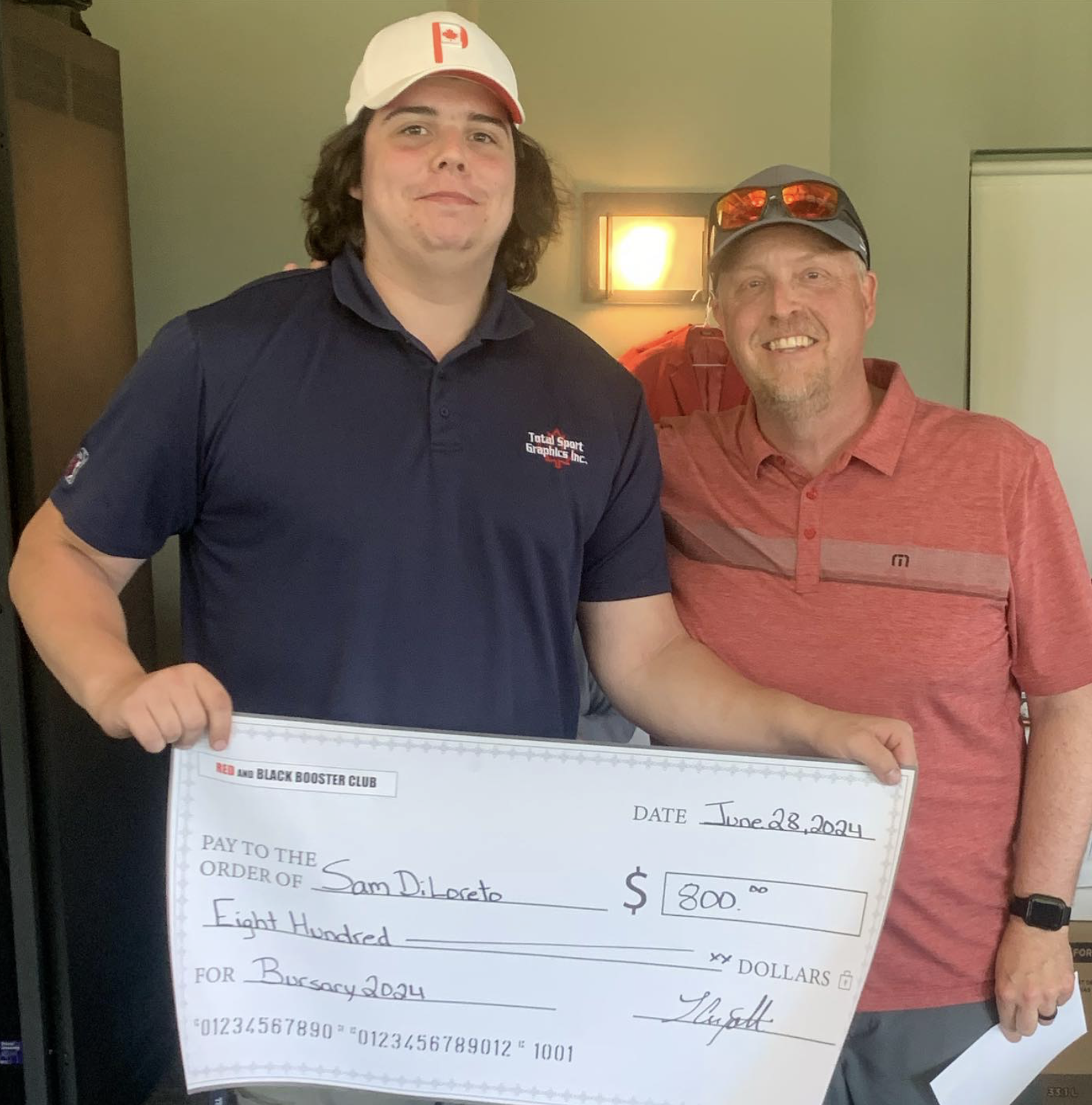 Two men standing indoors, holding a large check for $800 made out to Sam D. Letzo for the Bursary 2024, dated June 28, 2024. The man on the left is wearing a navy polo shirt with a logo that says Total Sport Graphics Inc. and a white cap with a red emblem. The man on the right is wearing a red polo shirt, sunglasses on his head, and a watch on his left wrist.