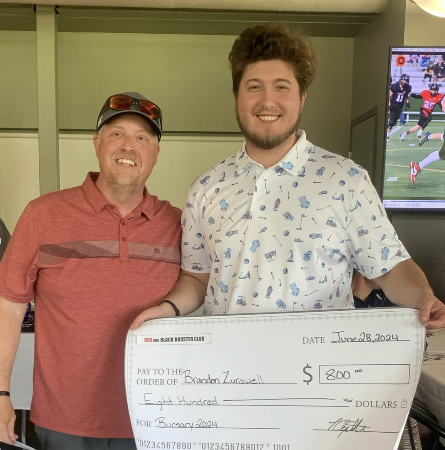 Two men smiling, one wearing a red polo shirt and sunglasses on his cap, the other wearing a white shirt with a sporty print, holding a large check made out to Brandon Zurawell for $800, dated June 28, 2024, in a room with a television showing a football game.