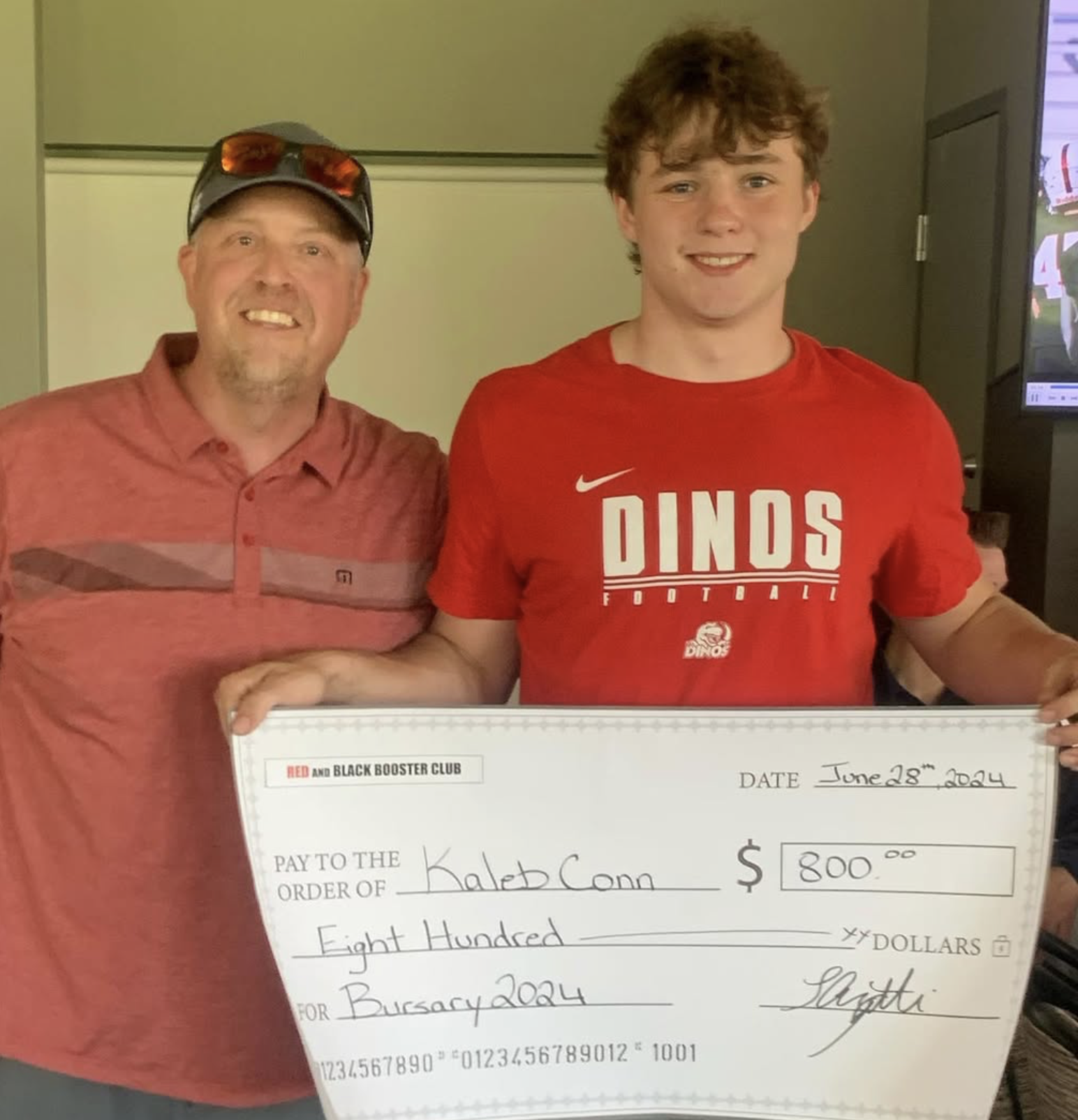 A young man in a red football shirt standing next to an older man wearing a cap and sunglasses on his head, both smiling, holding a large check made out for 800 dollars, dated June 28, 2024.