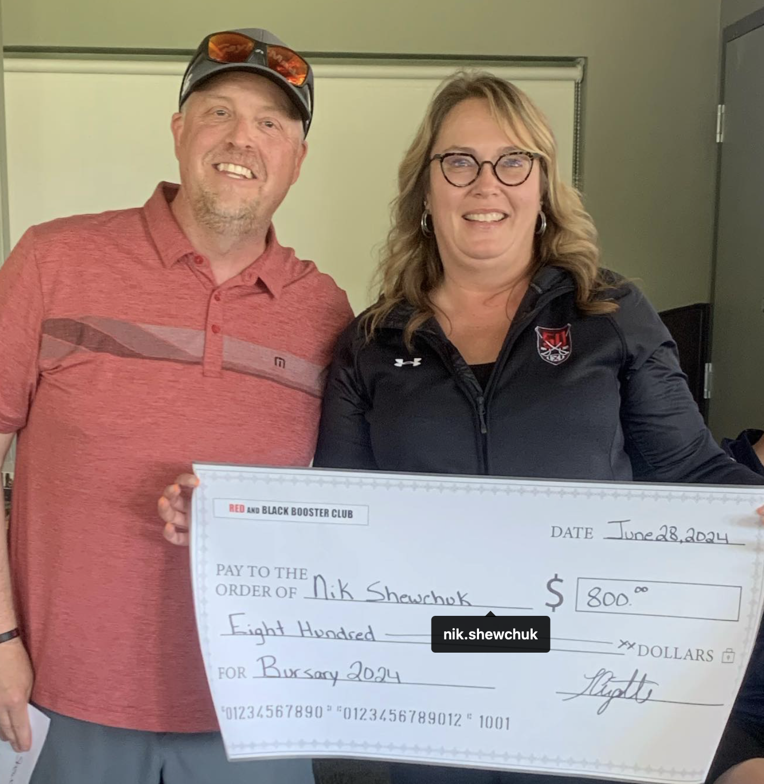 A man and woman smiling, holding a large check for $800 made out to Nik Shewchuk from the Red and Black Booster Club, dated June 28, 2024.
