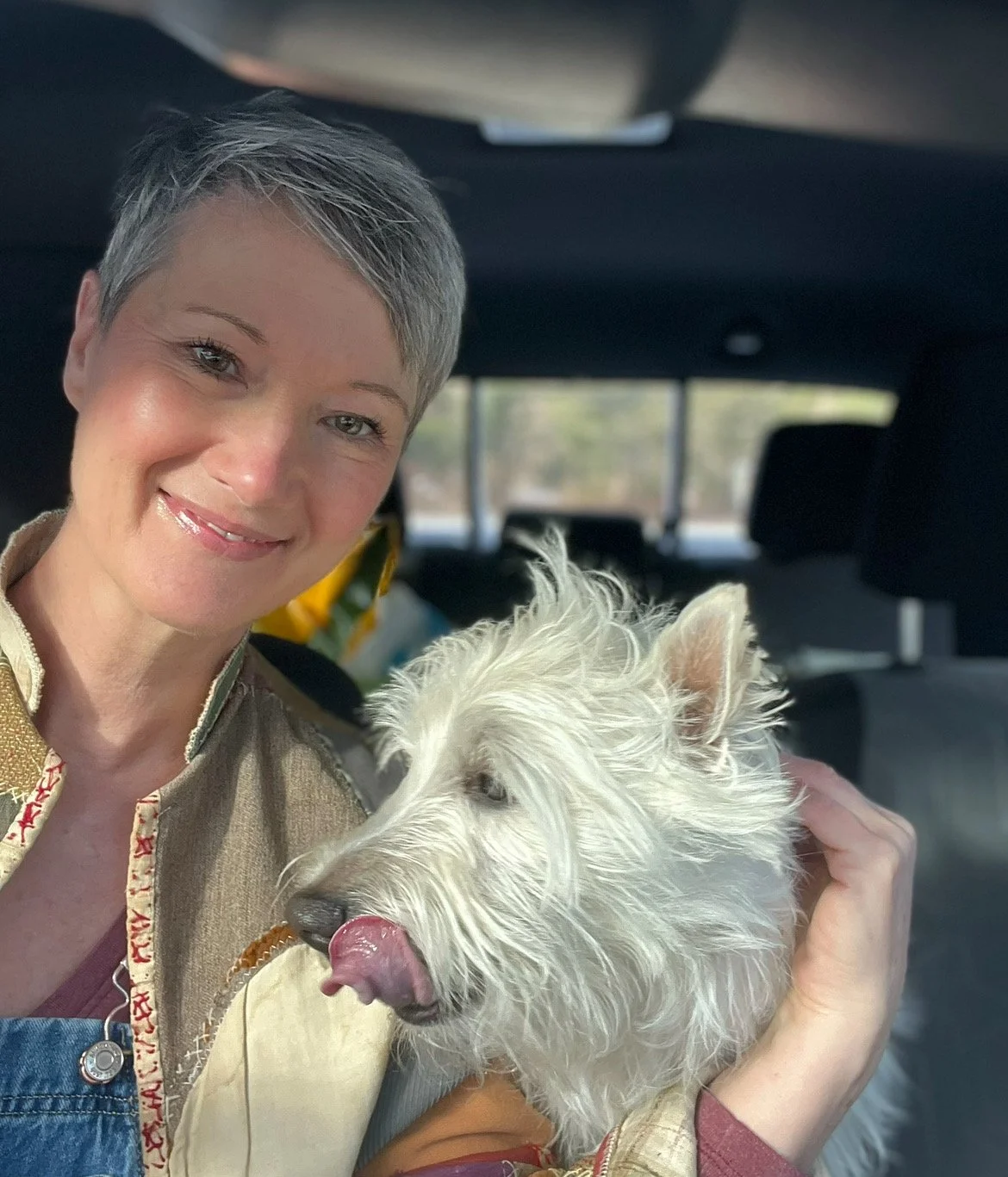 A woman with short gray hair smiling while holding a white West Highland White Terrier dog in a car.