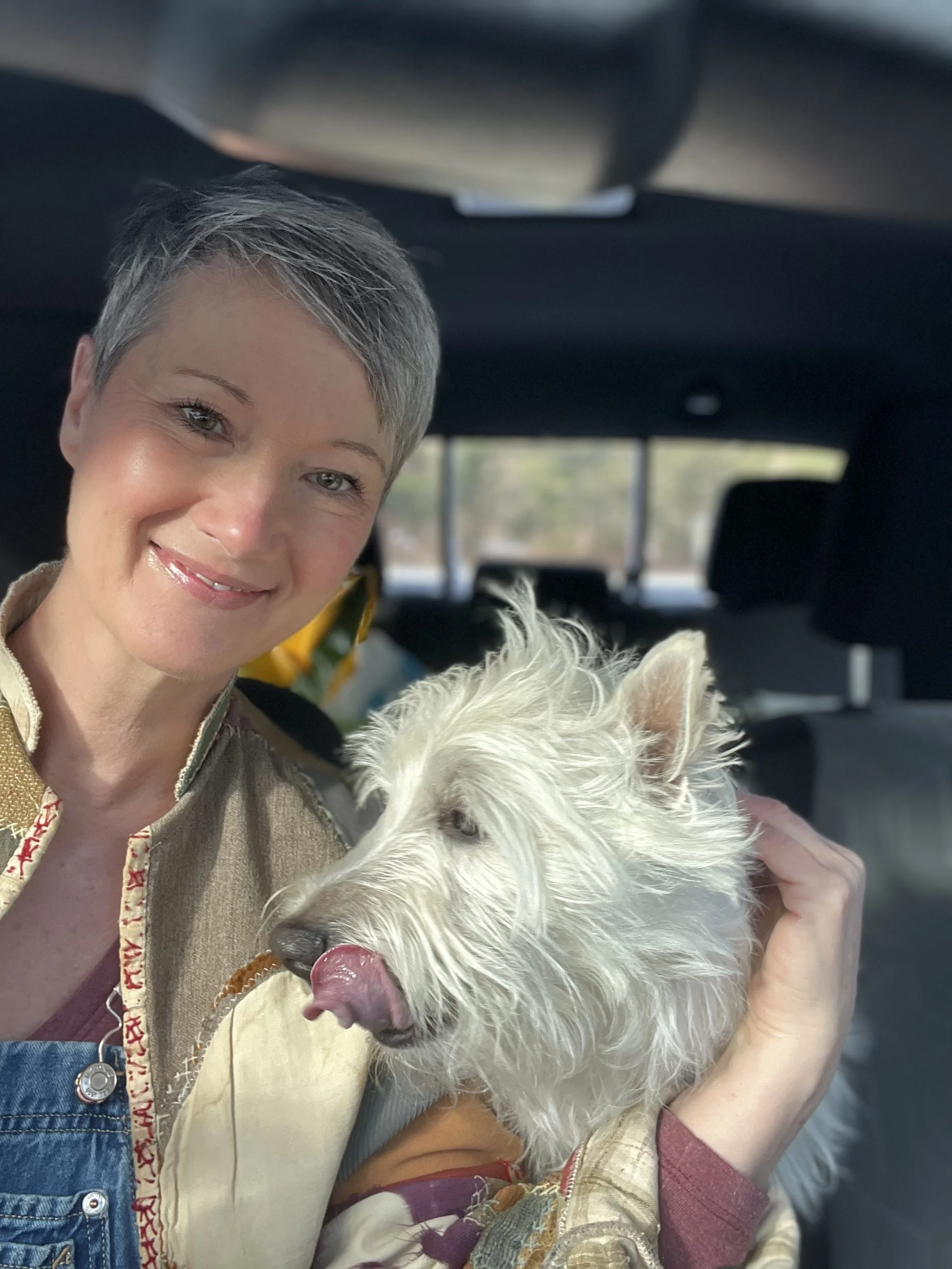 A person with short gray hair smiling while holding a white dog with a pink tongue inside a vehicle.