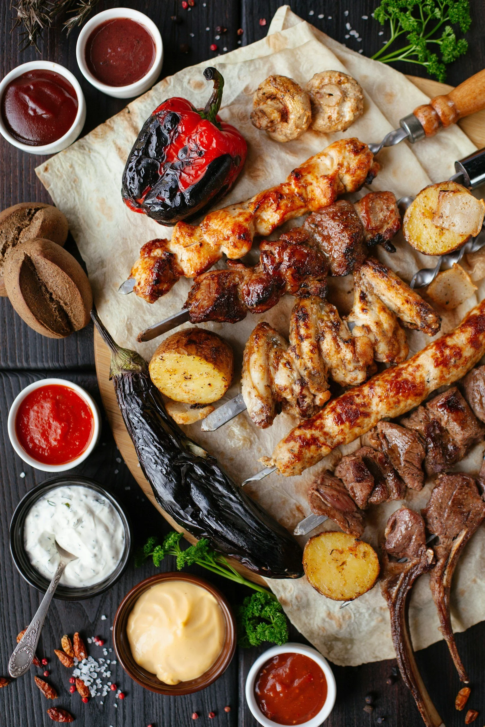 Assorted grilled meats on skewers with roasted potatoes, grilled peppers, and various sauces on a black wooden table.