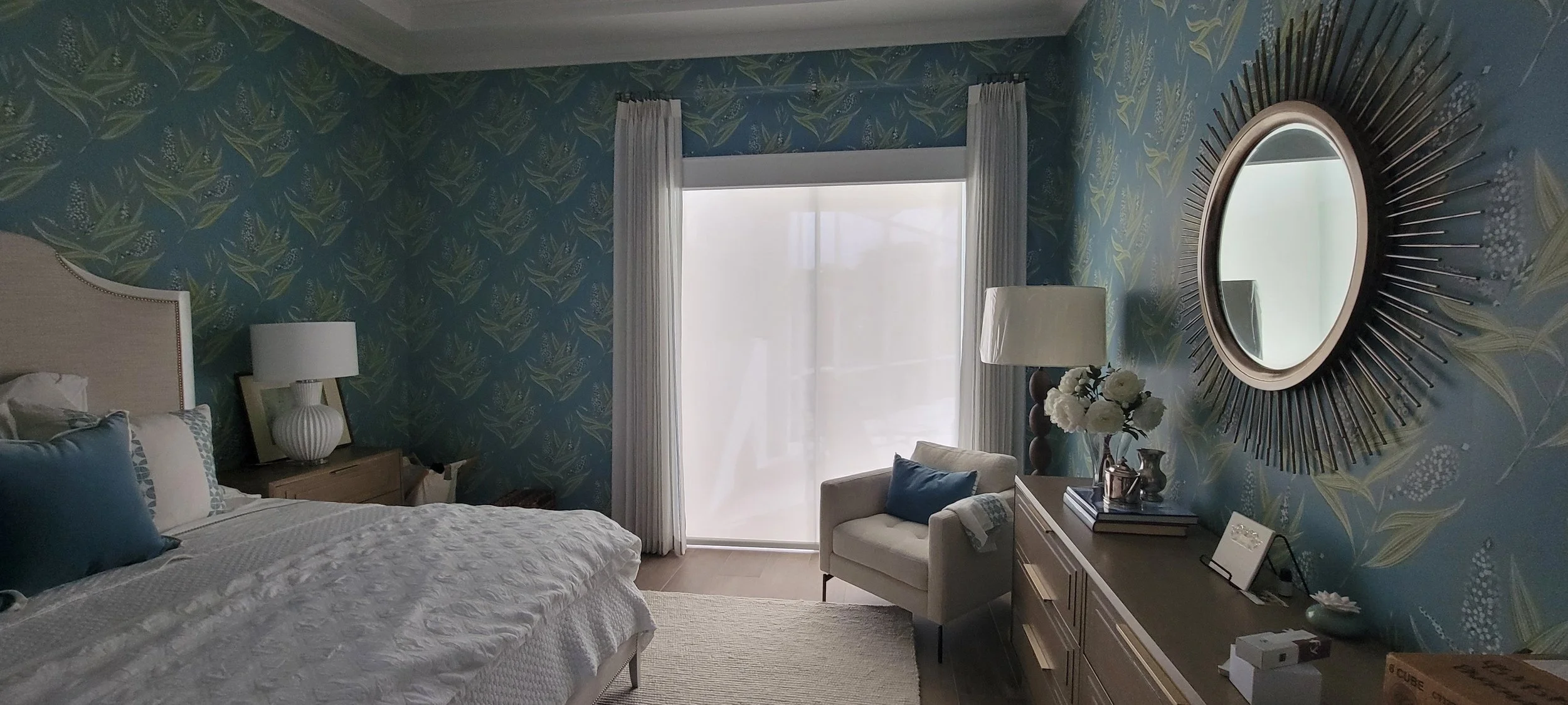 Stylish bedroom with blue floral wallpaper, white bedspread, circular mirror with radiating frame, white curtains, modern table lamp, armchair with cushion, dresser with decorative items, and large window.