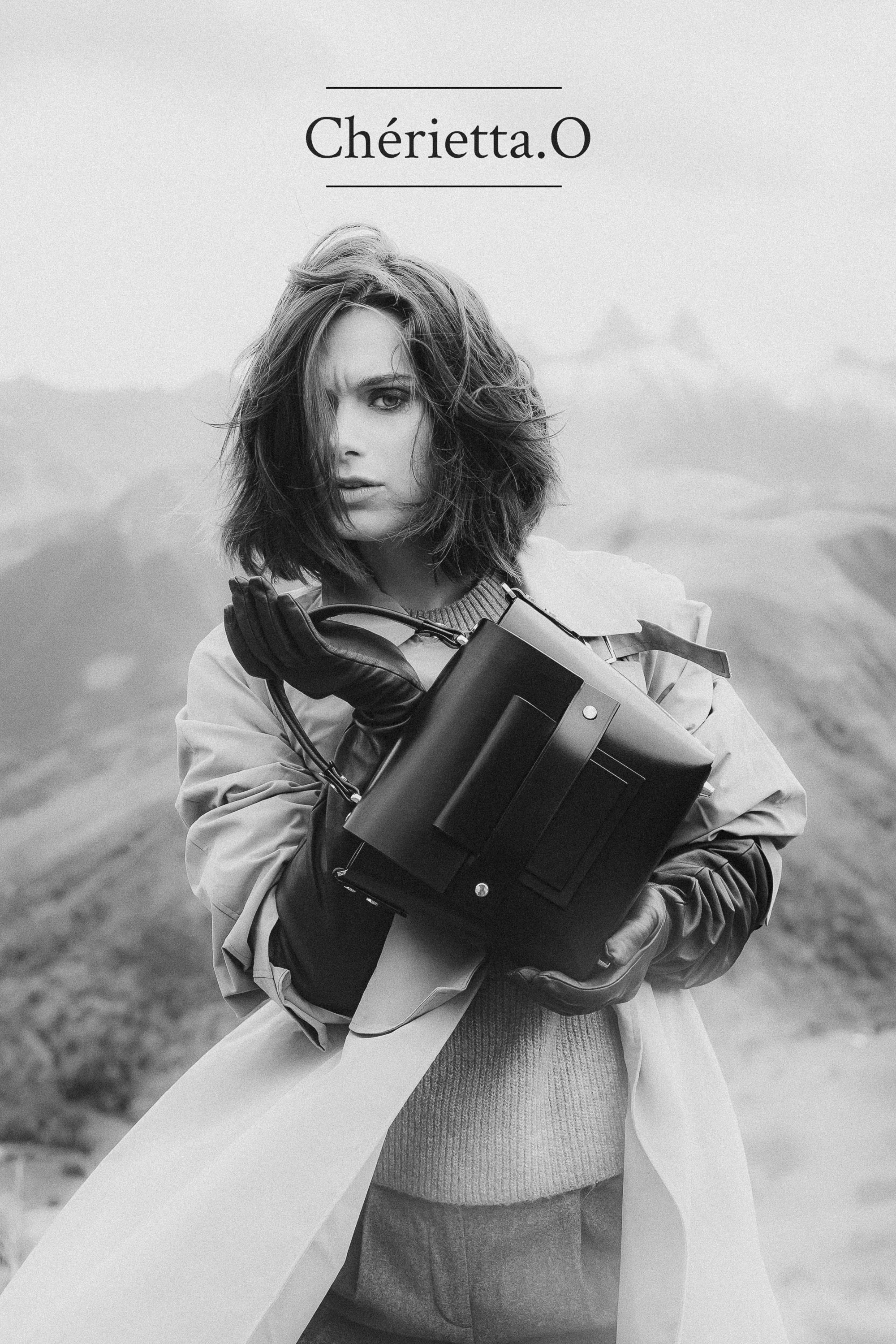 Portrait d'une femme en noir et blanc et tenant un sac en cuir dans un décor de montagne