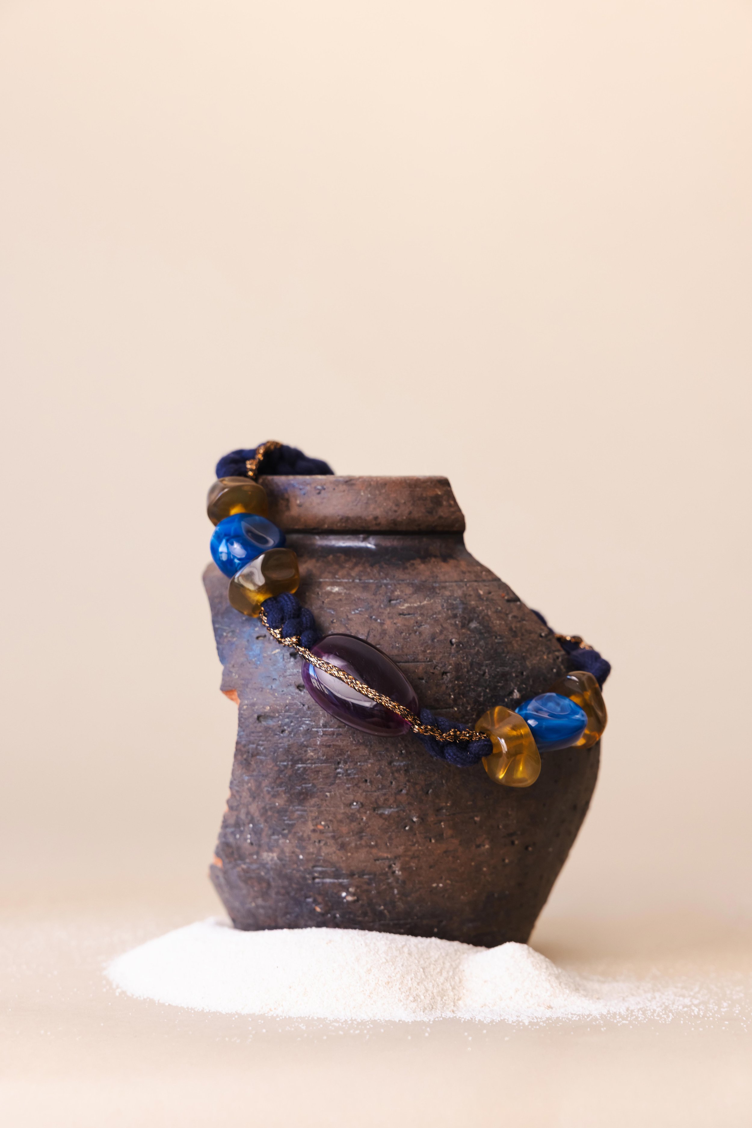 Nature morte d'un collier en grosses pierres bleues posé sur un morceau de poterie dans du sable