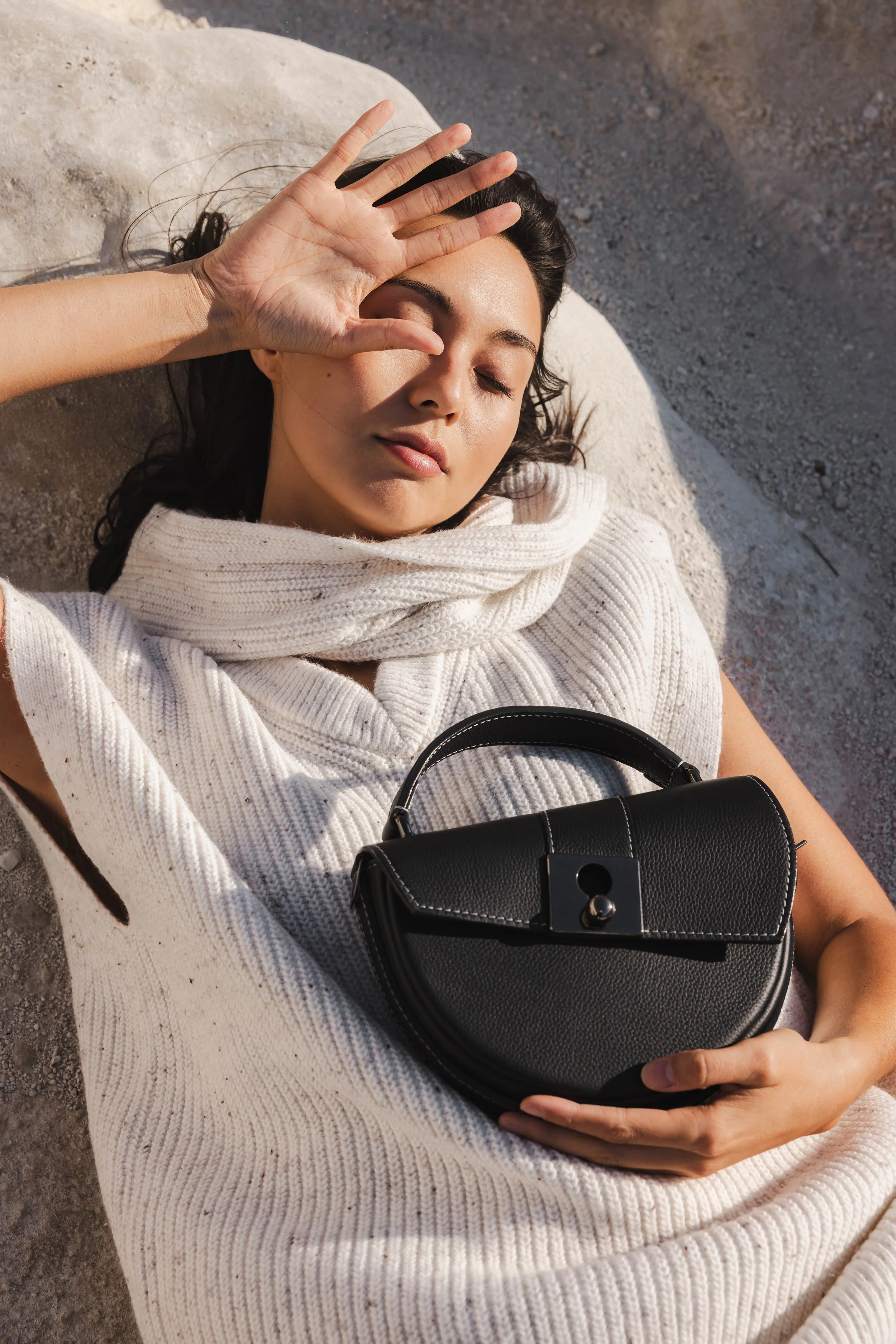 Portrait d'une mannequin en plein soleil, allongée sur une roche claire à Milos et tenant un sac en cuir noir