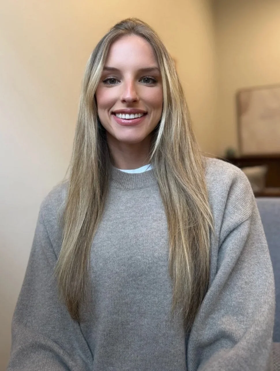Portrait of a smiling woman with long blonde hair, wearing a black shirt, against a light gray background.
