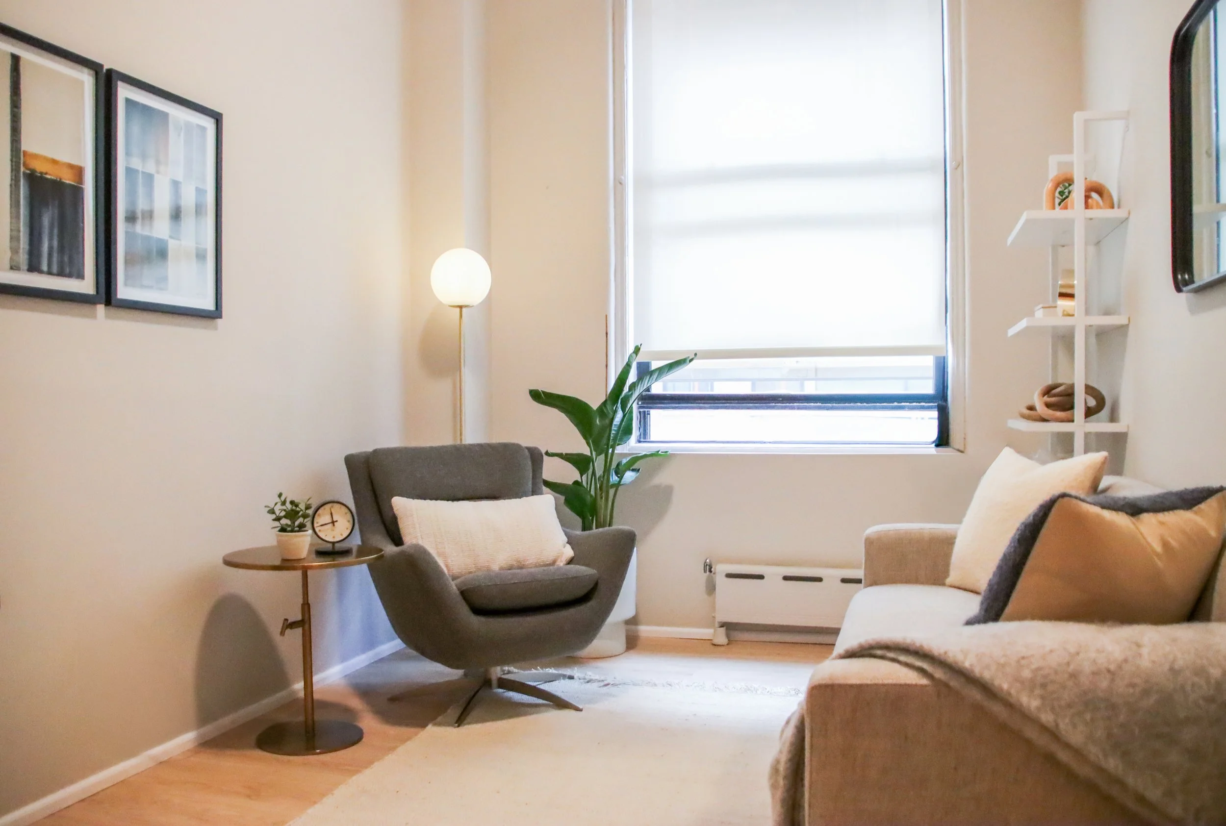A cozy living room with a beige sofa, beige rug, gray armchair with a pillow, a small round side table with a clock and plant, a tall floor lamp, a large green plant, and a window with a white rolled shade. There are three framed pictures on the wall and white shelves with decorative items.
