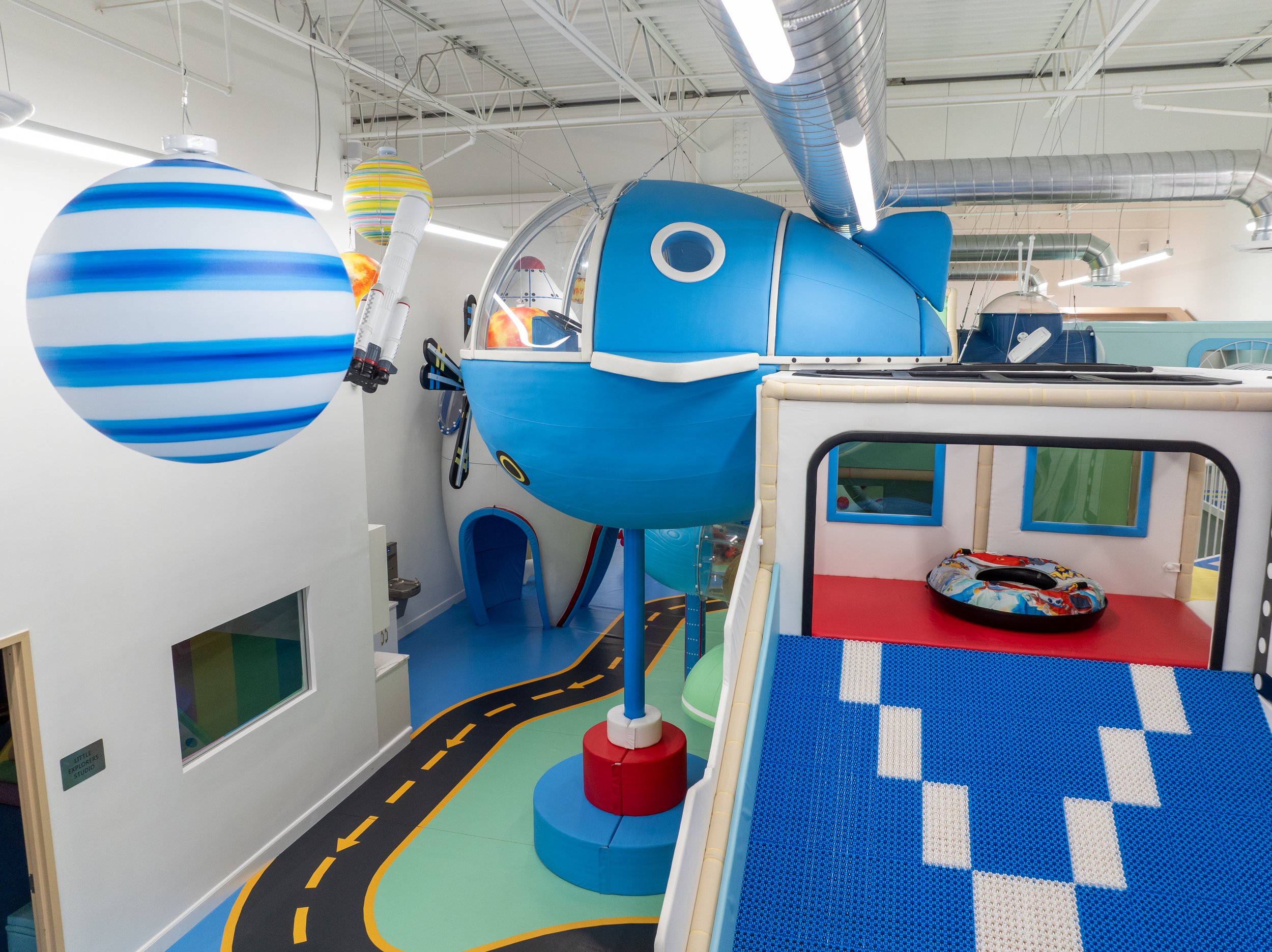 Indoor playground with astronaut-themed decorations, including a large blue spaceship, hanging planets, and a playhouse area with a small boat on a red surface.
