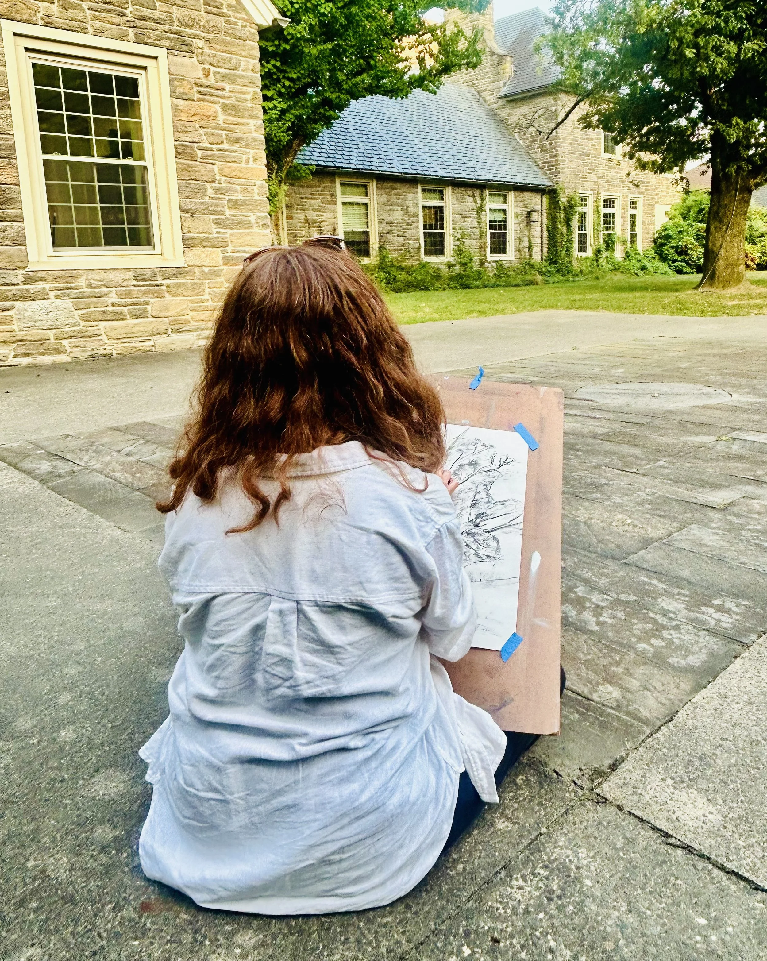 A person with long red hair sitting on the sidewalk, drawing or painting a tree on a sketchpad propped up on a board taped with blue painter's tape, outside a house with stone walls and large windows, surrounded by trees.