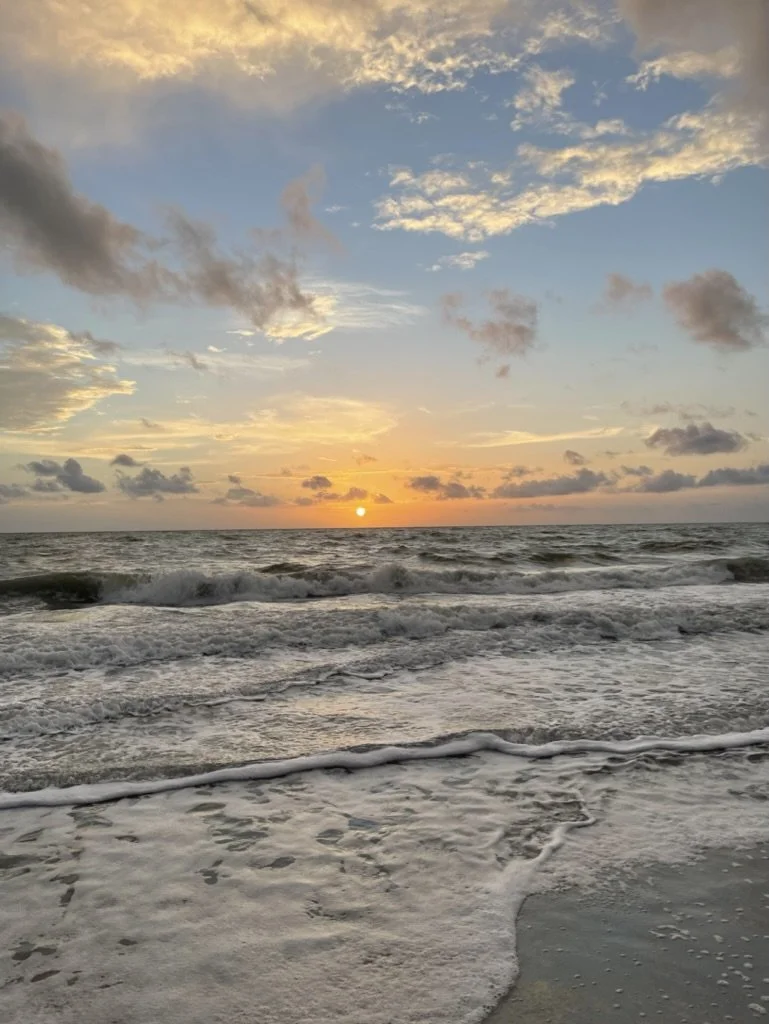 Sunset over the ocean with waves crashing onto the sandy beach, scattered clouds in the sky, and a peaceful, scenic view.