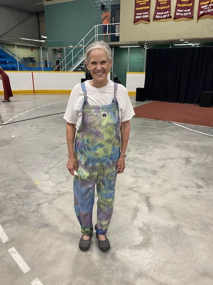 An older woman with gray hair smiling, standing at the Butterflies in Ballgowns market booth at the Rossland fall fair, wearing a white t-shirt and tie-dye butterflies in ballgowns overalls made sustainably from a bed sheet in Nelson BC