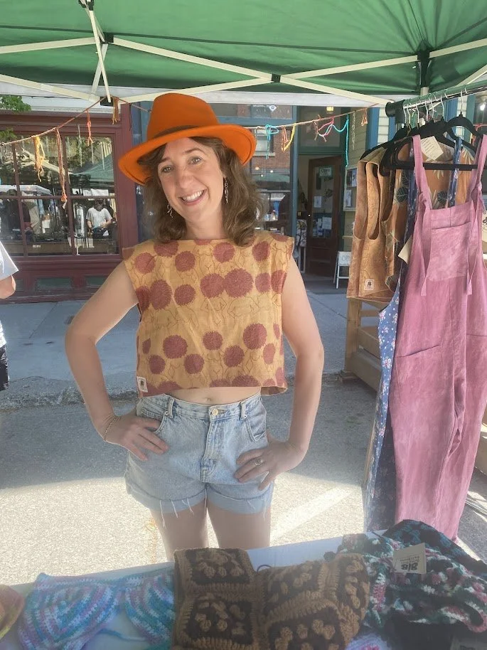 Young woman in sleeveless crop top with dahlia flower pattern- made sustainably by Butterflies in Ballgowns using natural fibres and dyes - denim shorts, orange hat, standing at Butterflies in Ballgown market stall at the Nelson Farmers Market.