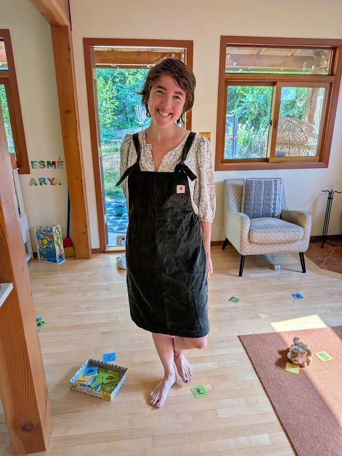 A smiling woman with short brown hair standing barefoot in a living room, wearing a green corduroy Butterflies in Ballgowns dress, made in Nelson BC, from upcycled fabric