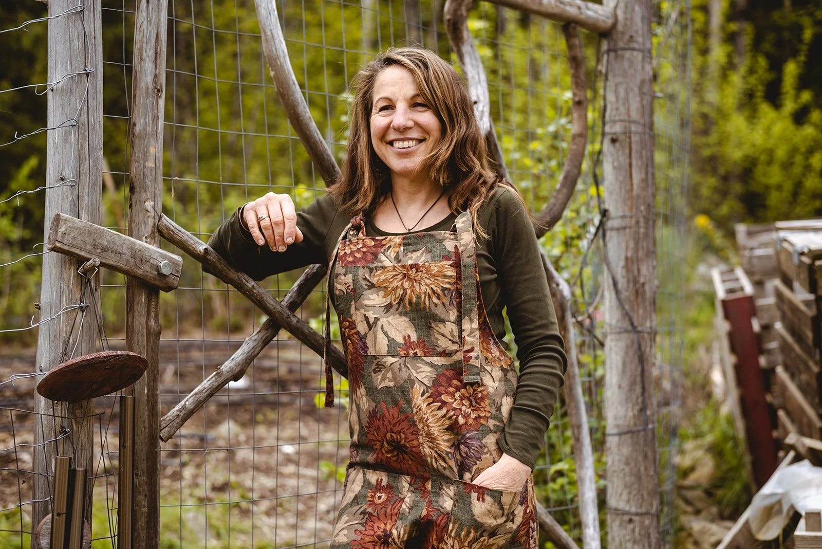 A woman smiling and standing outdoors in front of a wire fence, wearing a floral pair of Butterflies in Ballgowns dungarees, made sustainably from upcycled fabric in Nelson BC 