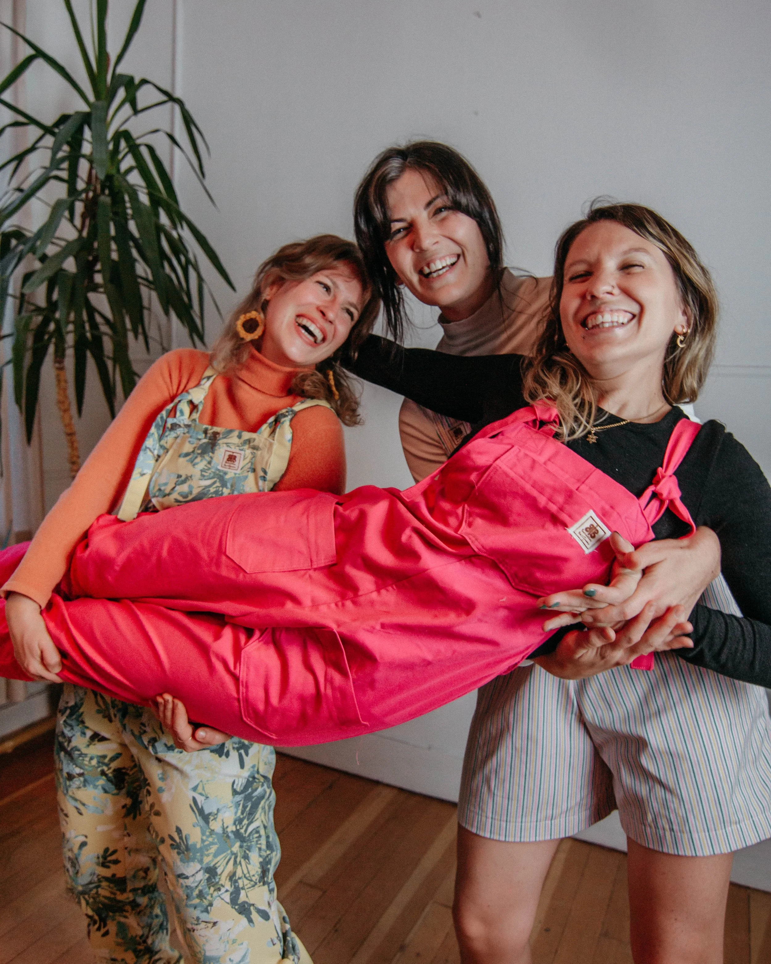 3 women all wearing Butterflies in Ballgowns dungarees. Two of them are holding one of them up, and they are all smiling and laughing.